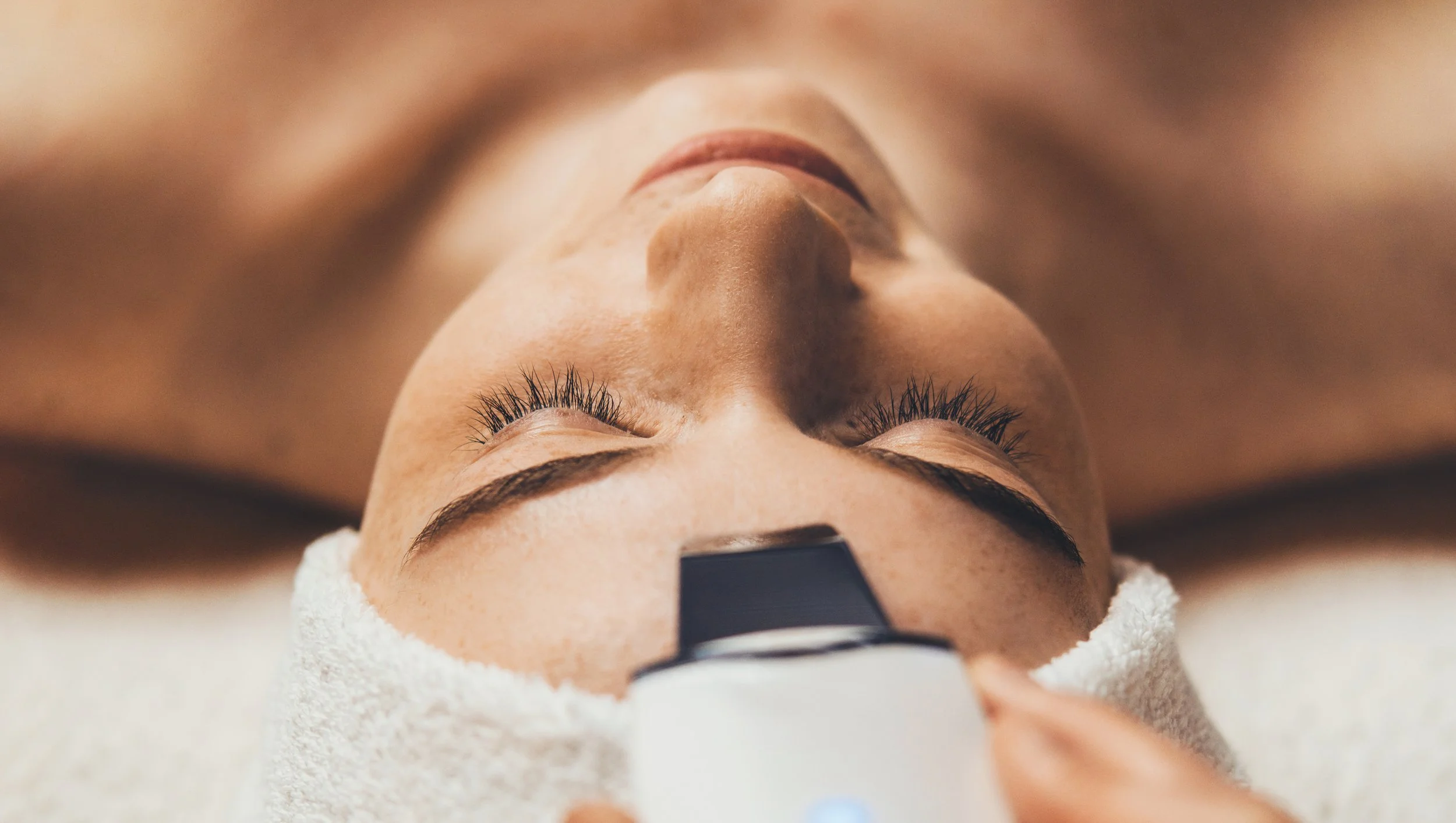 Close-up of a woman lying on a treatment bed with her eyes closed, receiving a facial or beauty treatment, with a white towel around her head and face.
