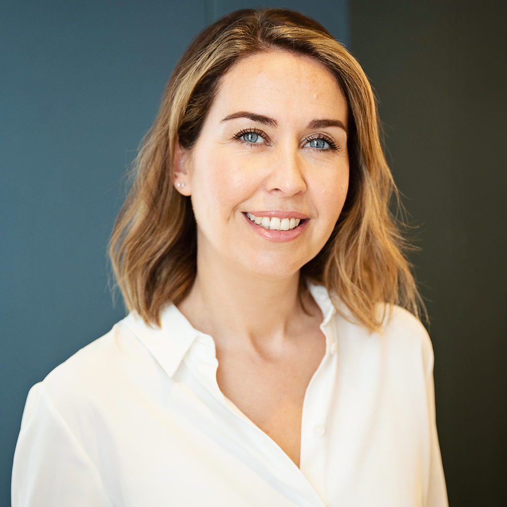 A woman with shoulder-length light brown hair, blue eyes, and a friendly smile, wearing a white blouse against a dark background.
