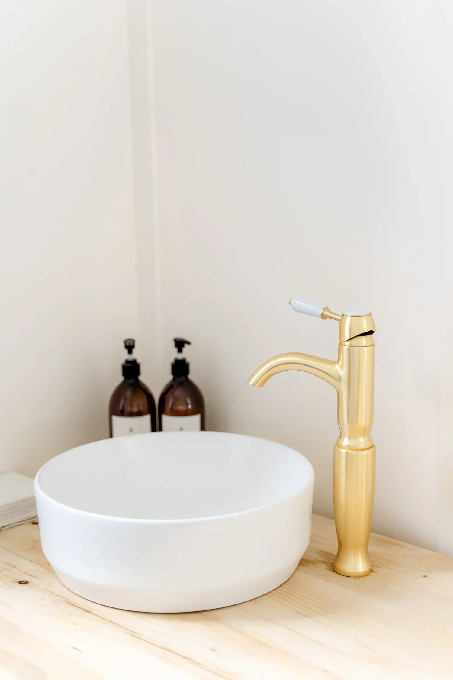 A white circular vessel sink on a wooden countertop with a gold faucet. In the background, two brown soap dispensers and two black faucets are visible against a plain white wall.