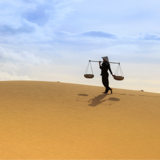 A person in desert clothing carrying a traditional balance scale, walking across golden sand dunes under a partly cloudy sky.