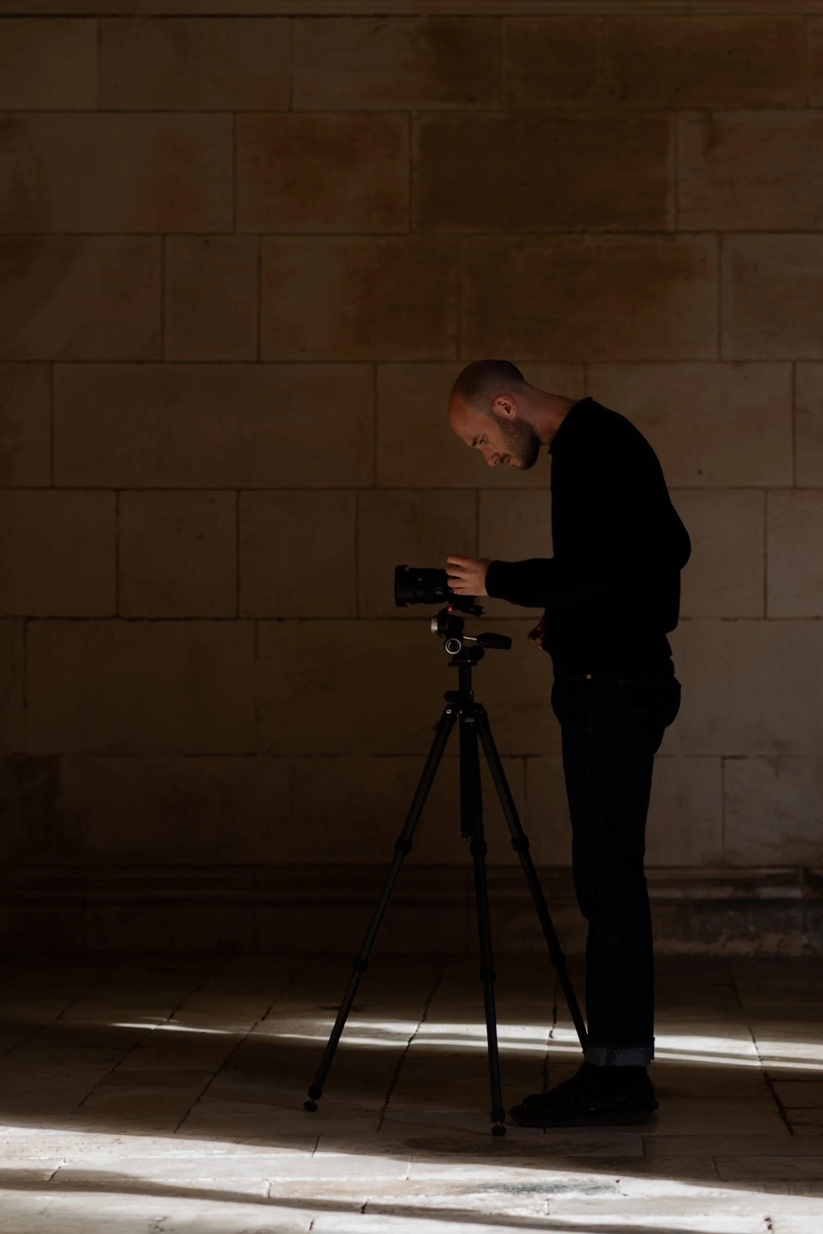 A man with a shaved head and beard wearing a black jacket and pants, standing behind a camera on a tripod, in front of a textured wall with lighting creating shadows.