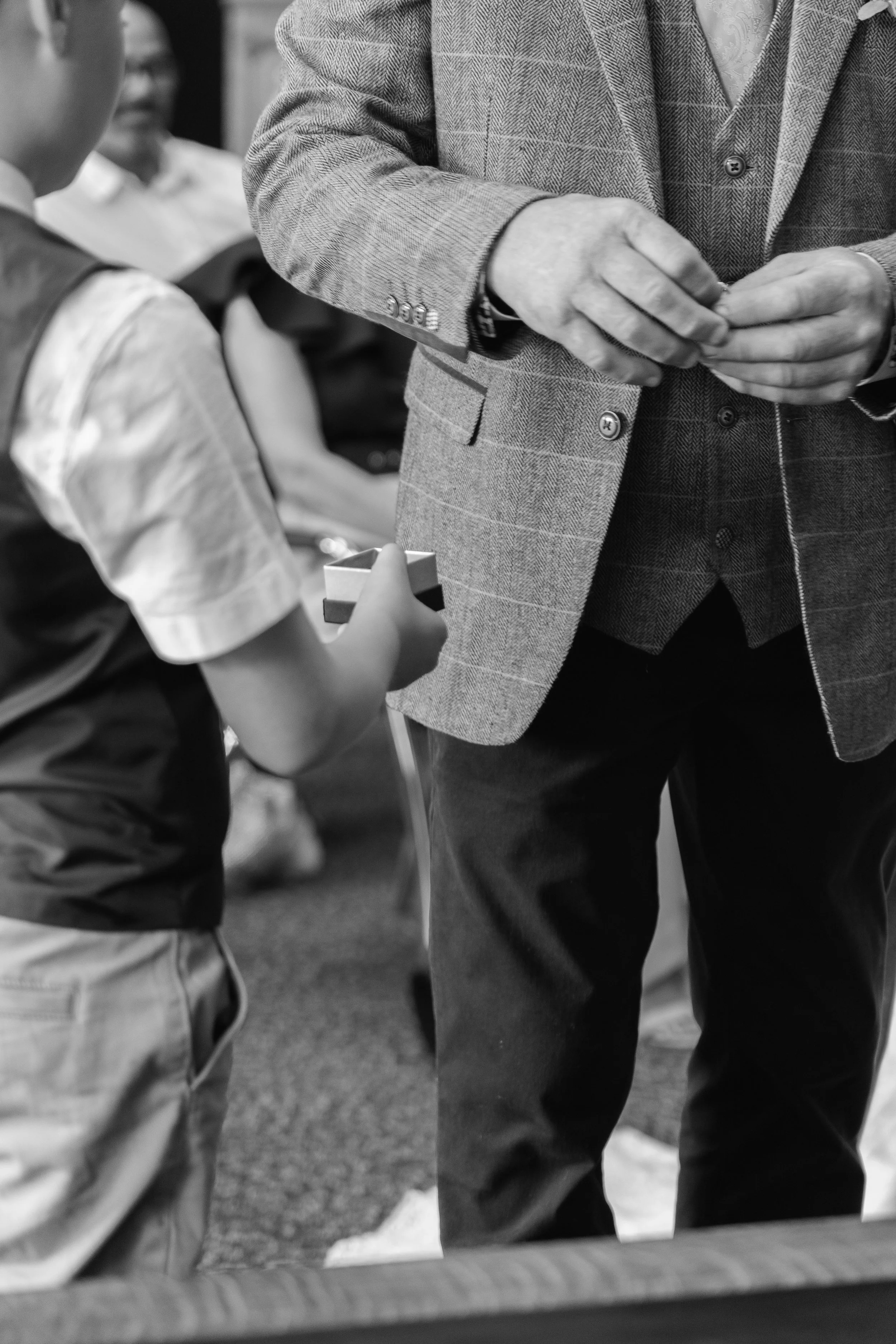 A person wearing a patterned blazer is handing something to a young boy, who is holding a small box, in a social setting with other people visible in the background.