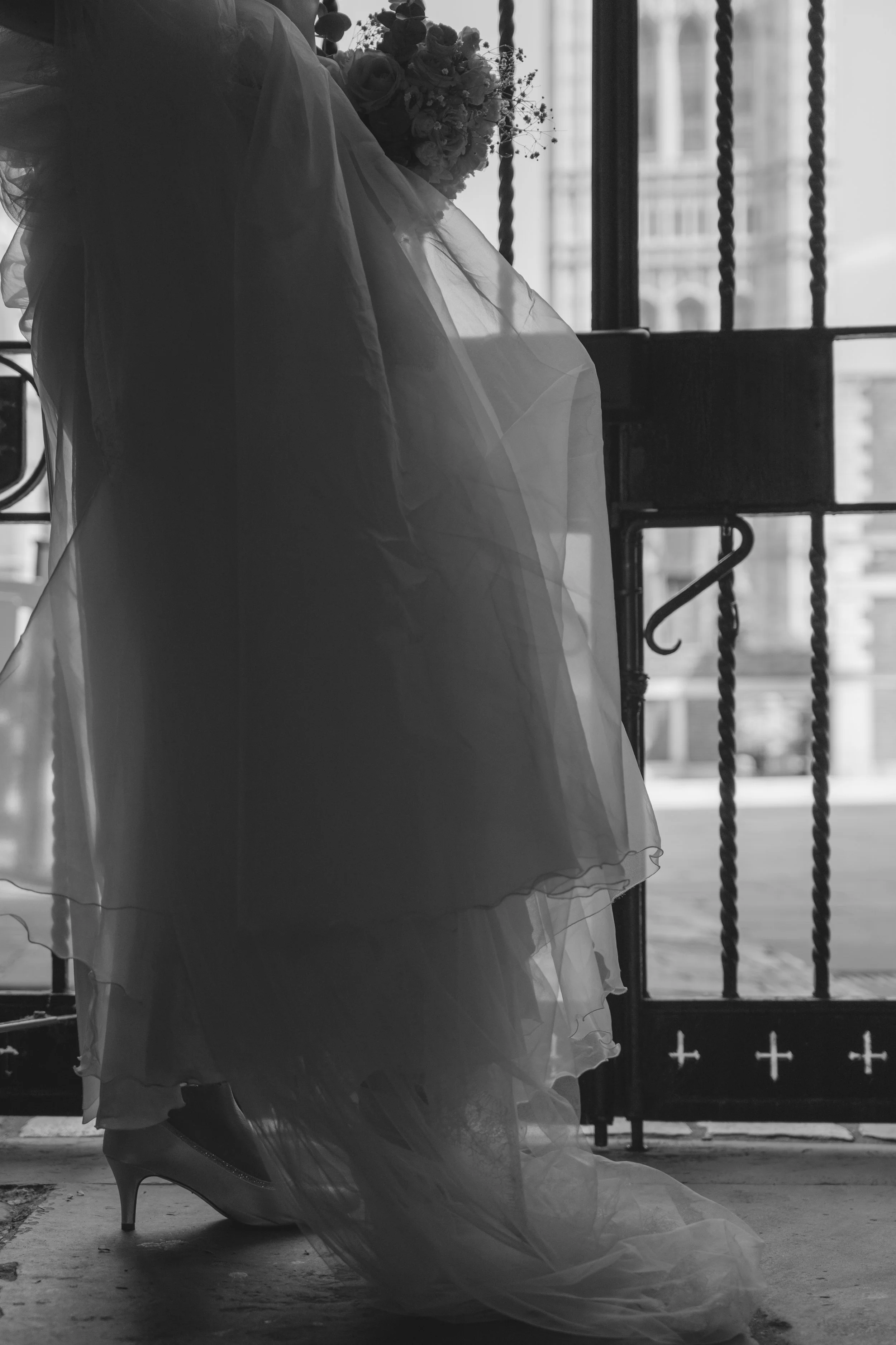 Black and white photo of a woman standing by a barred window, wearing high heels and a voluminous dress, holding a bouquet of flowers.