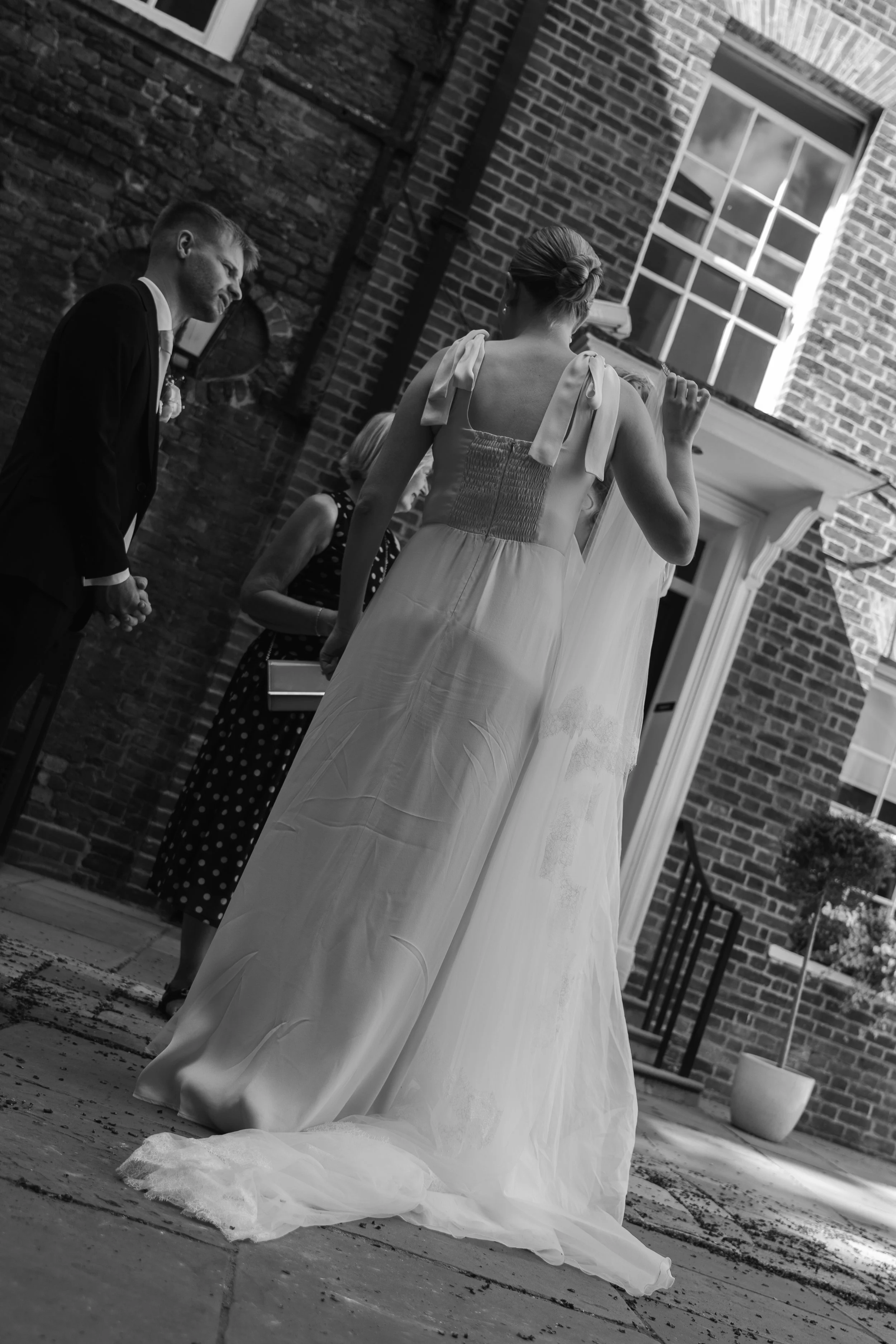A bride in a wedding dress with tied shoulder straps, standing outdoors on a brick patio in front of a brick building with large window. A man in a suit stands nearby, and two women, one in a polka dot dress, are in the background.