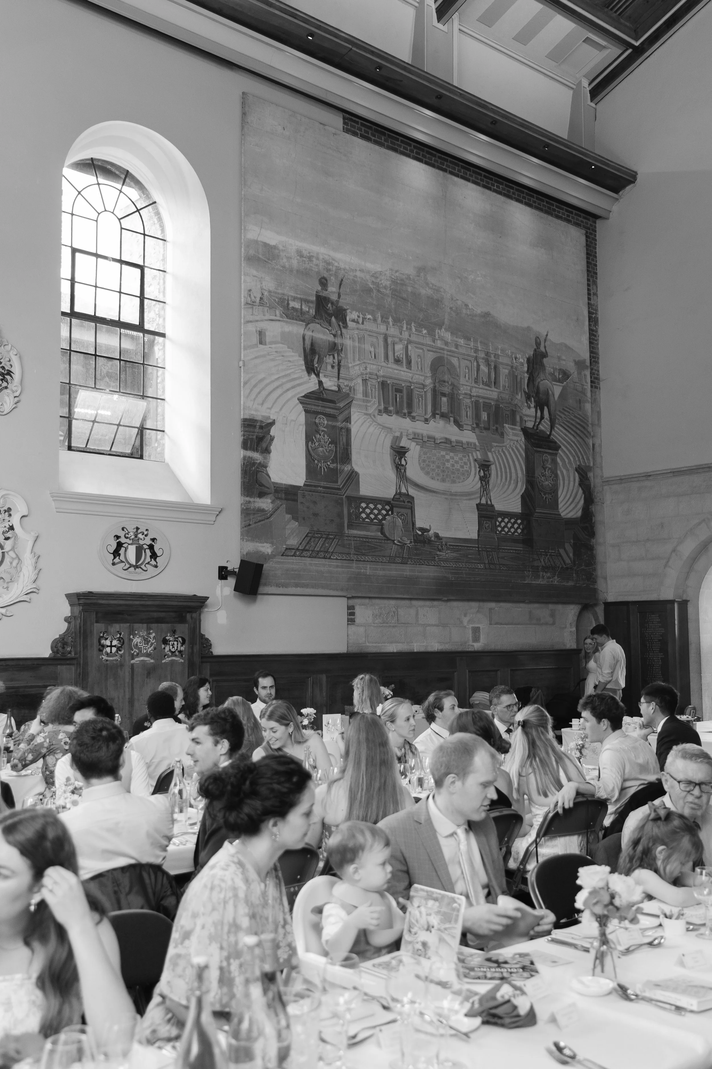 A large group of people seated at tables during a formal event or celebration inside a historic building with large arched windows and artwork depicting historical figures on the wall.