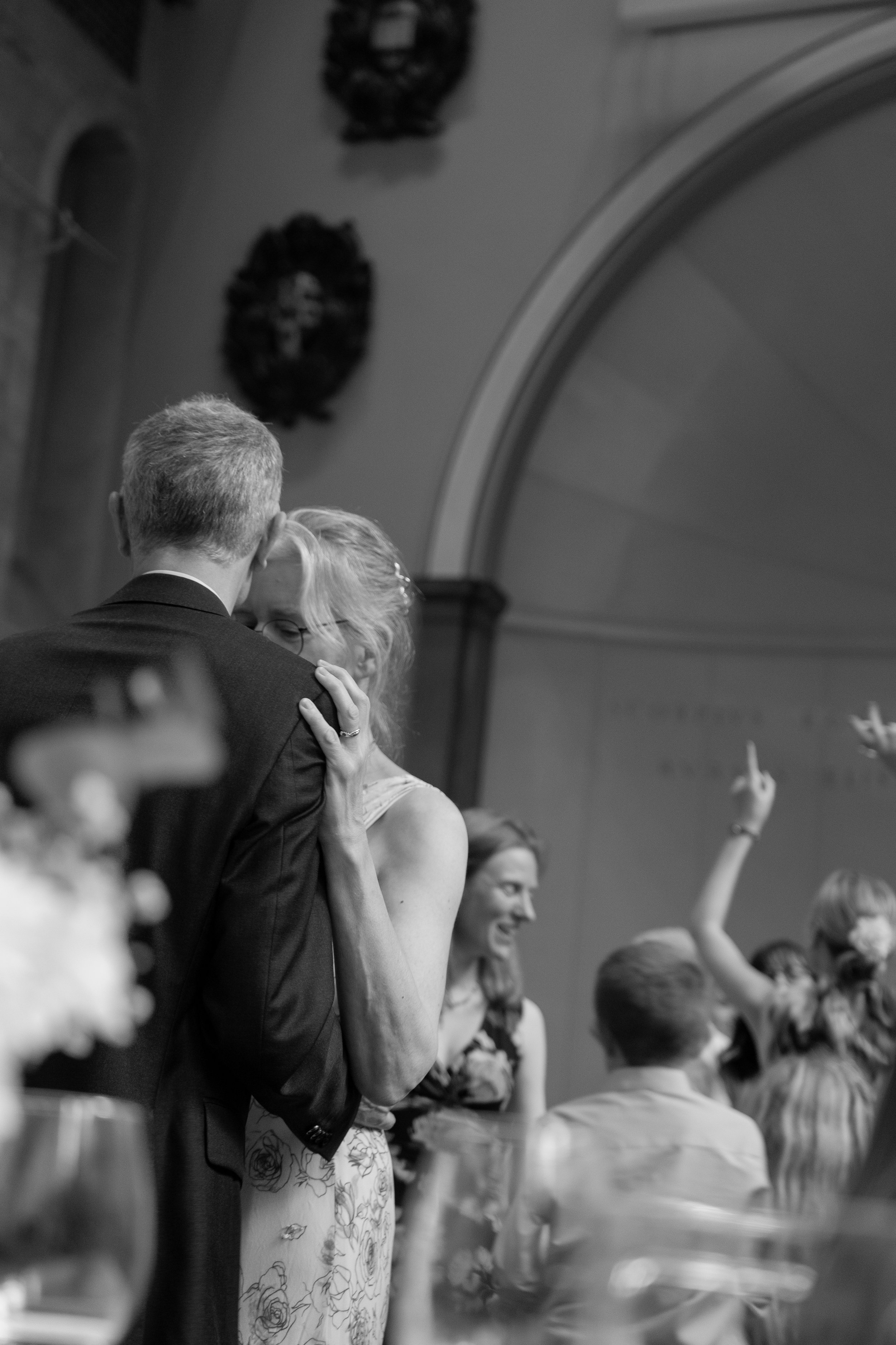 A black-and-white photo of a wedding reception showing a couple dancing closely, surrounded by seated and standing guests, with some raising their hands or taking photos in the background.