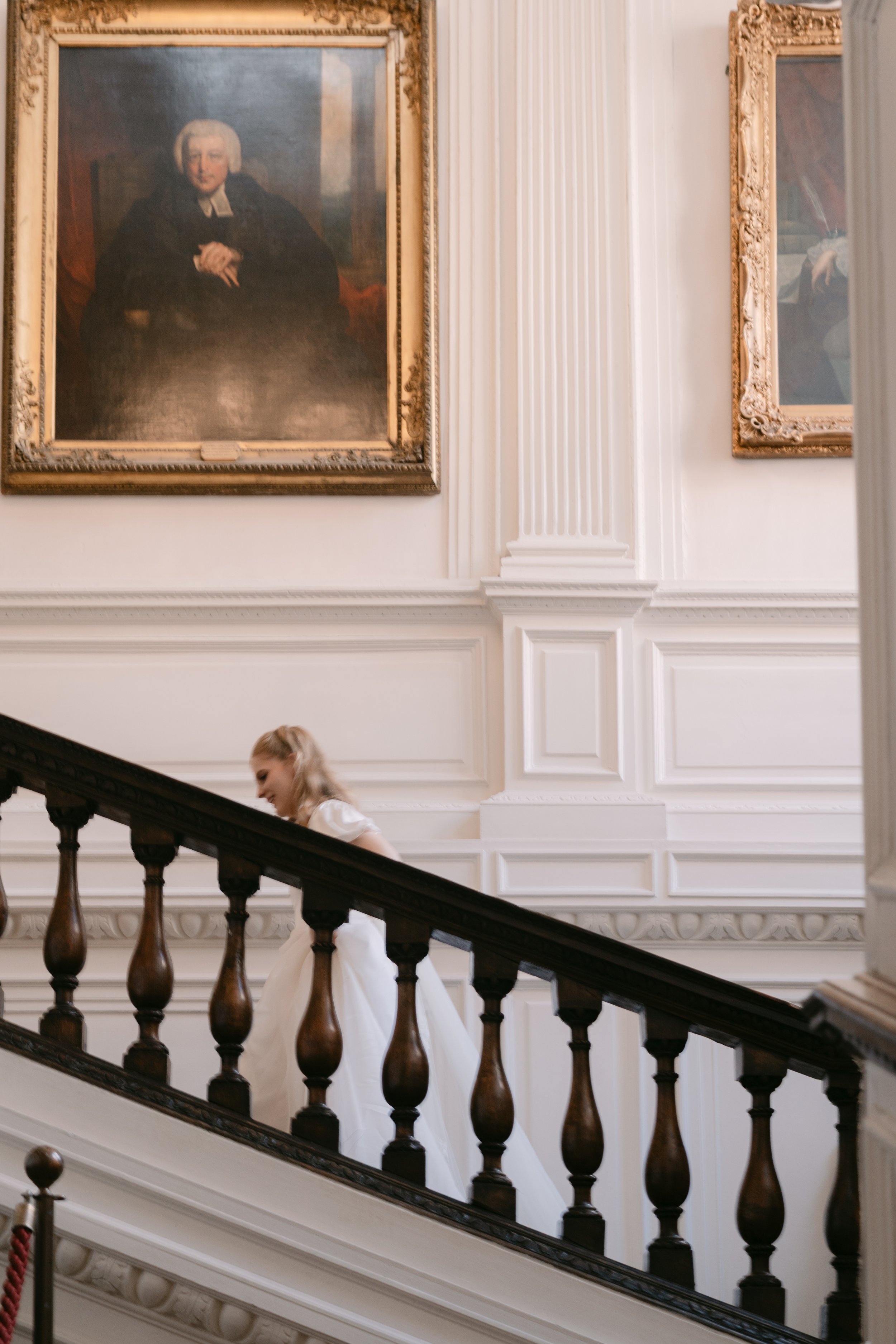 A young girl in a white dress walking down a staircase in a mansion or historic building with ornate white wall moldings and large framed paintings.