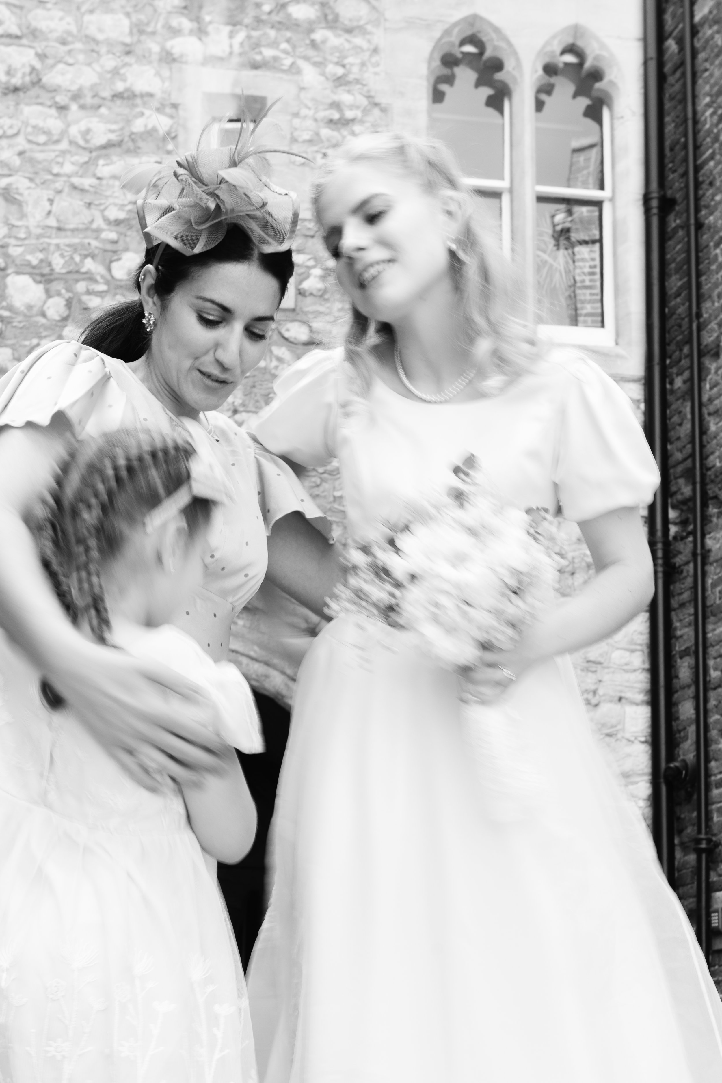 Two women in vintage dresses, one wearing a large hat with a bow, sharing a moment outdoors with a stone wall and Gothic-style windows behind them.