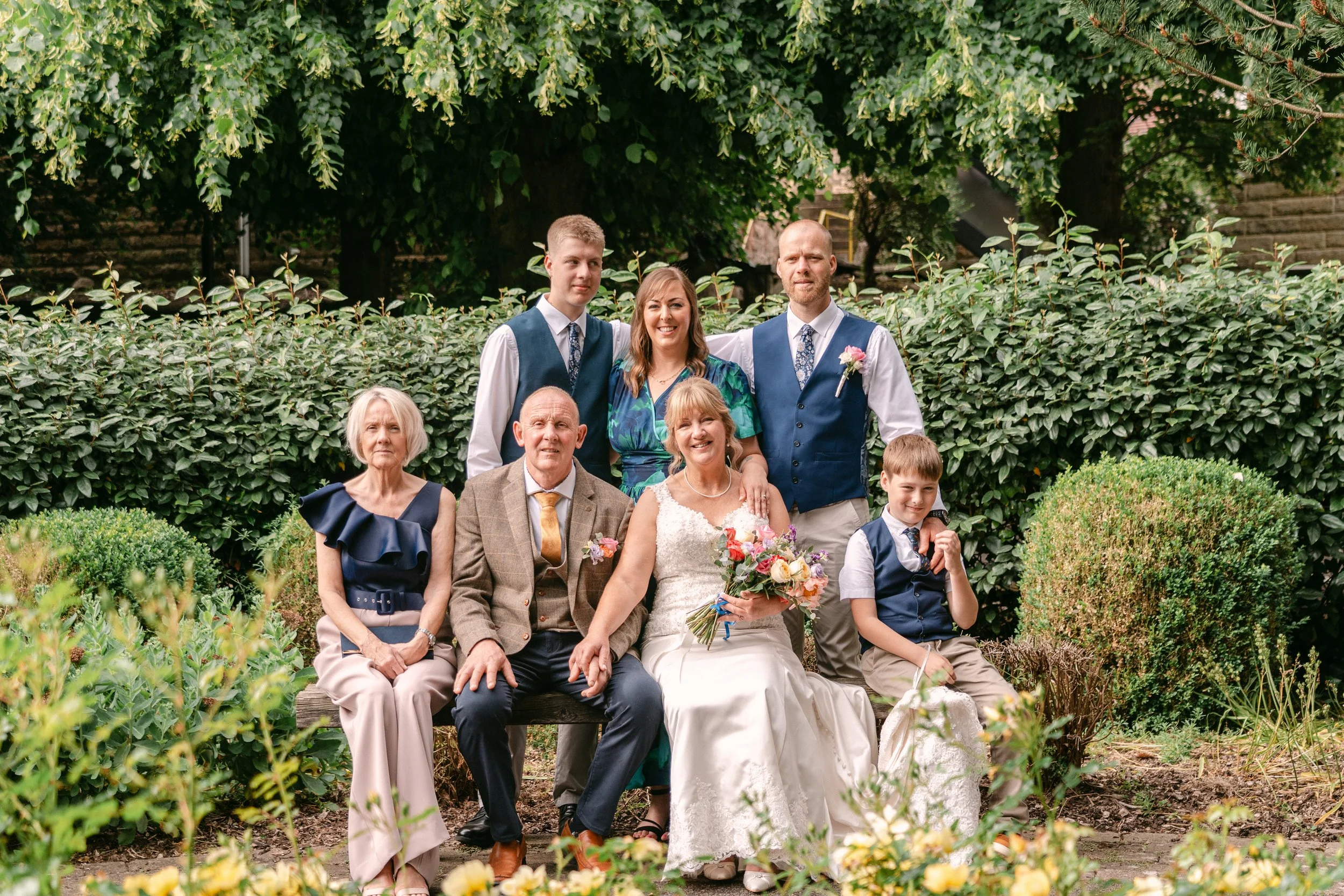 A wedding photo of six people sitting and standing in a garden with lush greenery and bushes. The bride in a white dress holds a bouquet, surrounded by five others dressed in formal and semi-formal attire.