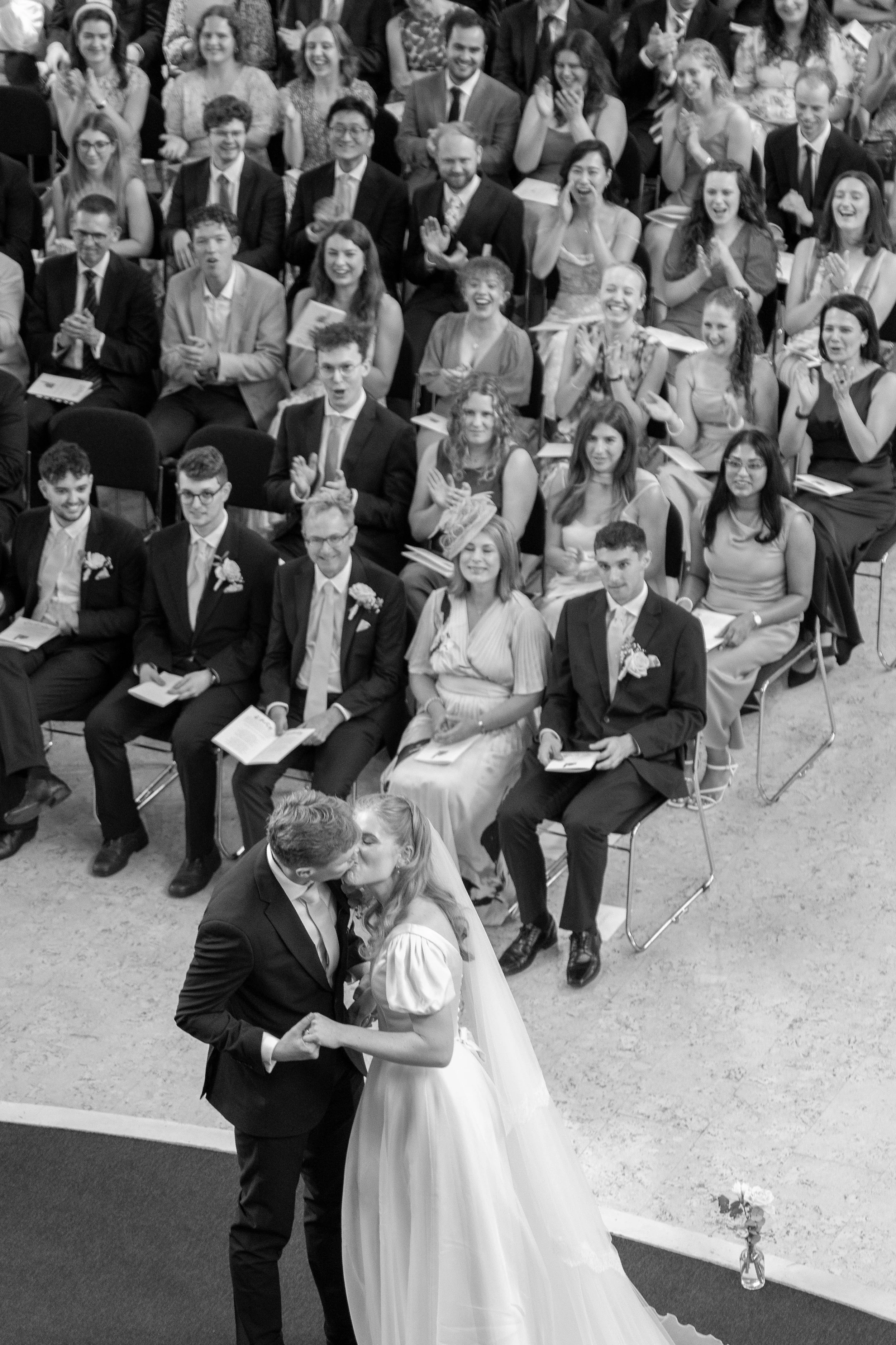 A bride and groom kissing and holding hands during their wedding ceremony in front of a seated audience, with guests clapping and smiling.