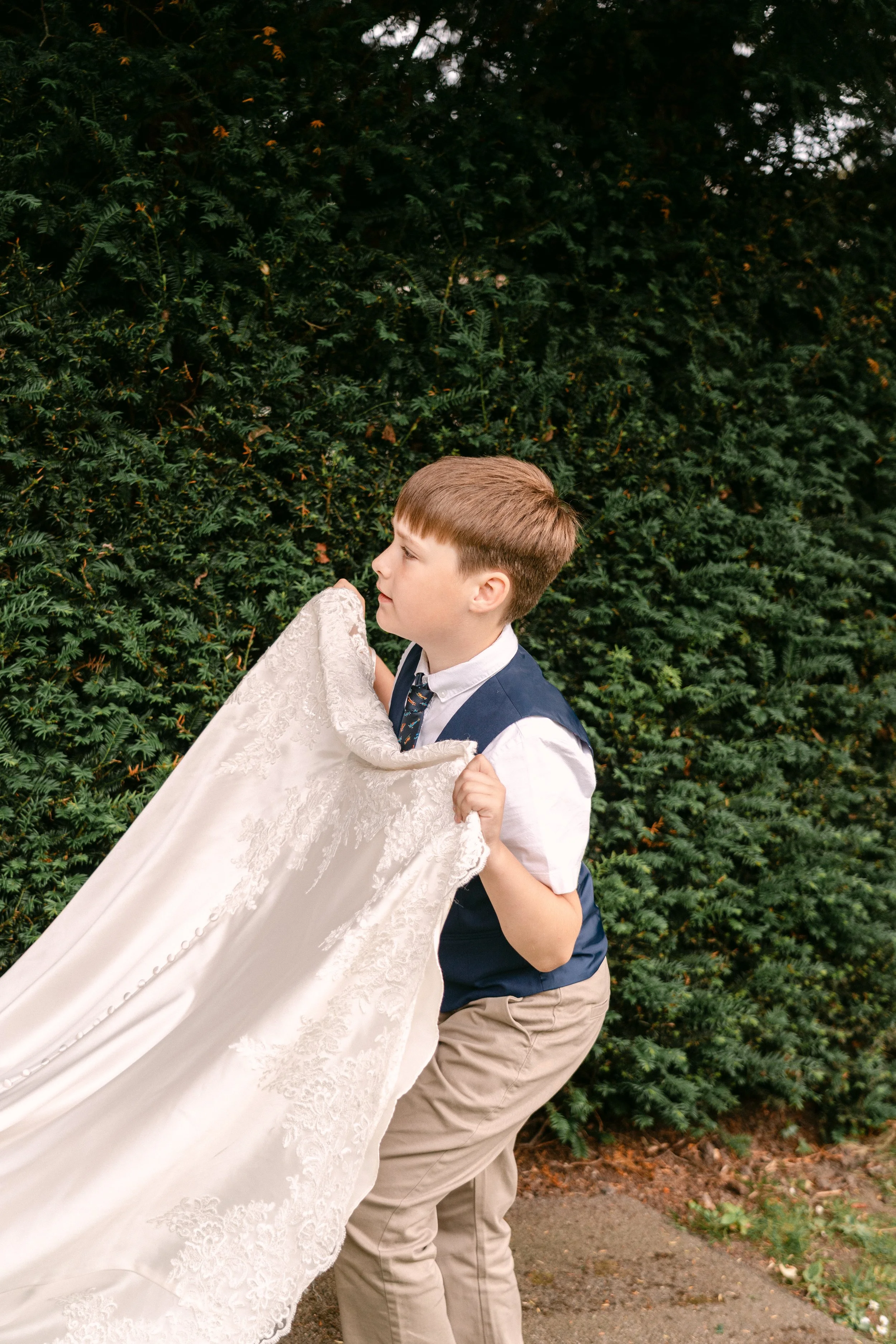 A young boy in a blue vest and khaki pants holds up a white, lace wedding dress in front of a green hedge in an outdoor setting.