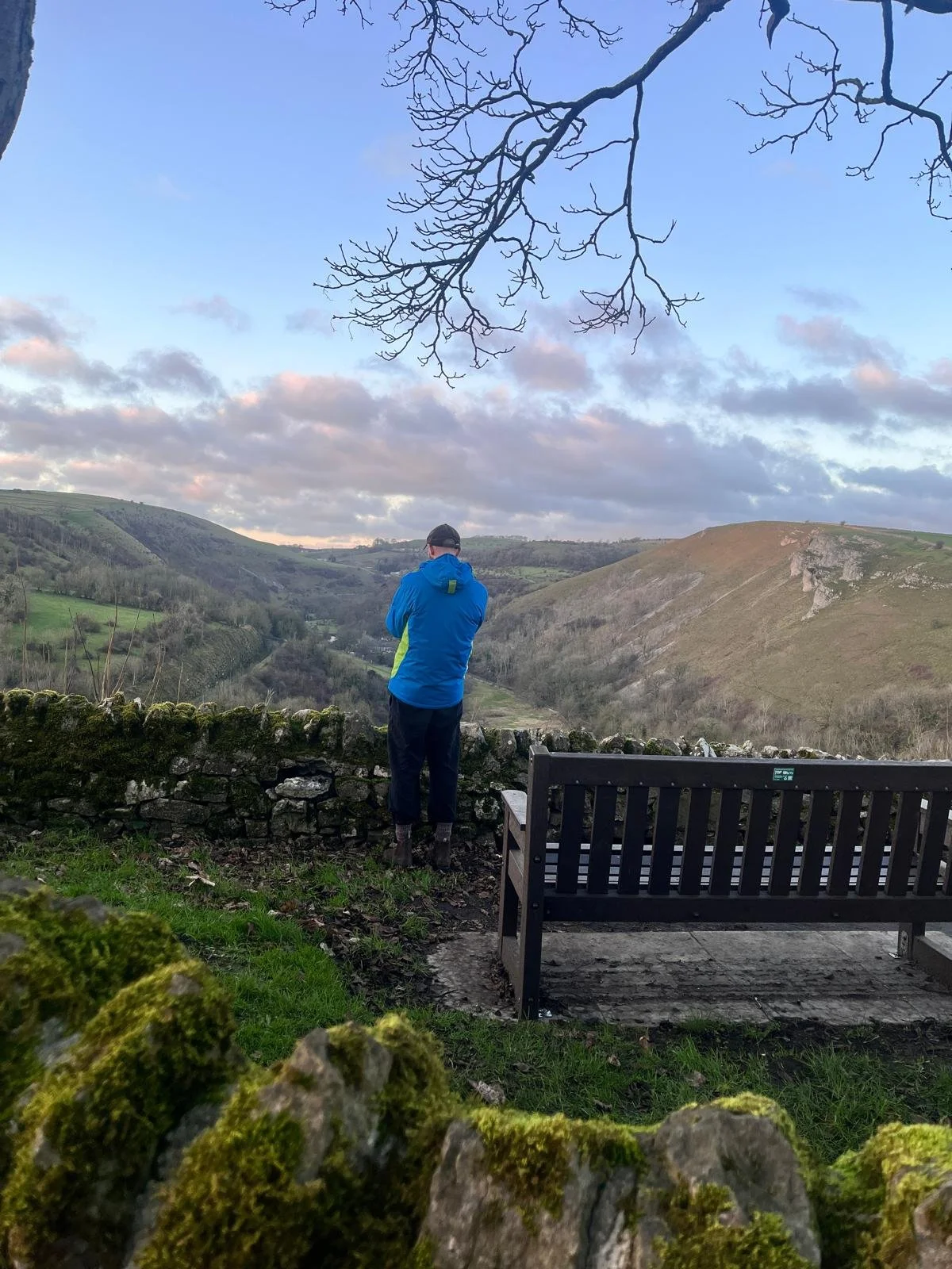 A person in a blue jacket and black pants stands overlooking a valley with hills and trees, next to a stone wall and a wooden bench, under a sky with scattered clouds.