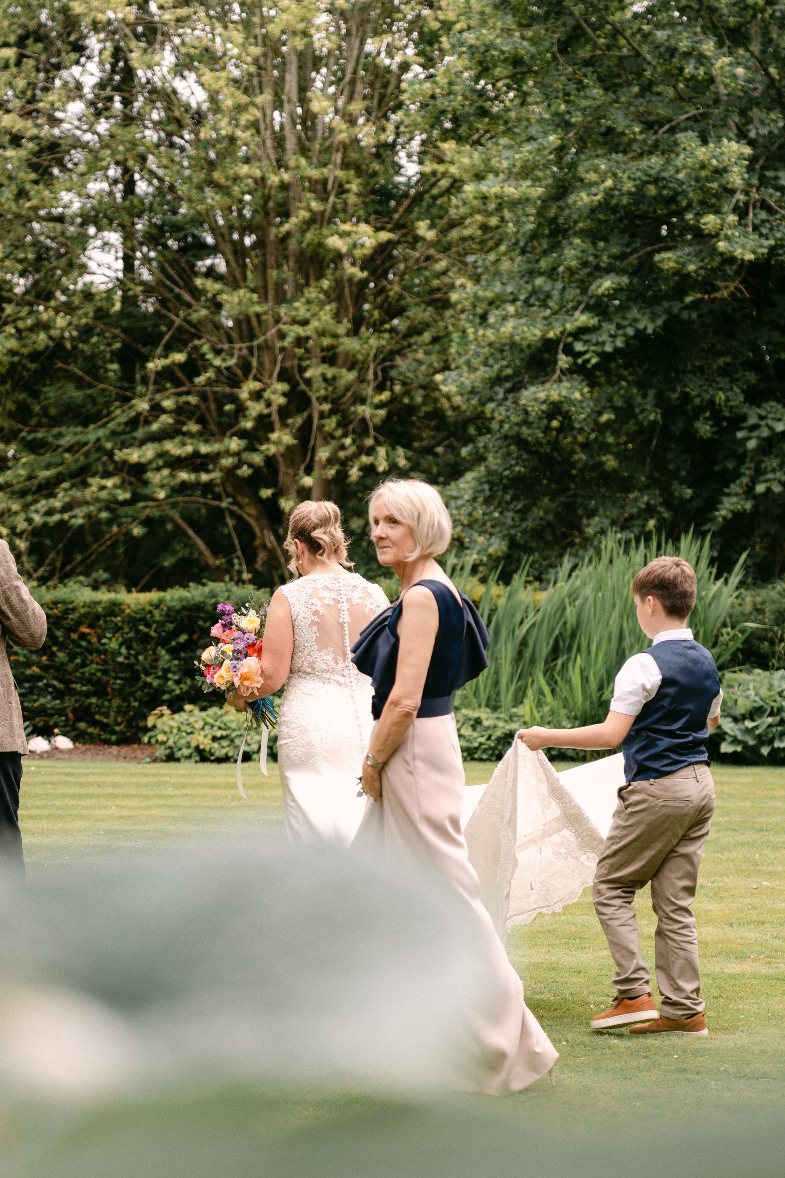 A woman in a white lace wedding dress holding a bouquet of flowers, accompanied by an older woman in a navy top and light-colored skirt, and a young boy in a vest and trousers, standing on a manicured lawn with trees in the background, likely at a wedding ceremony.