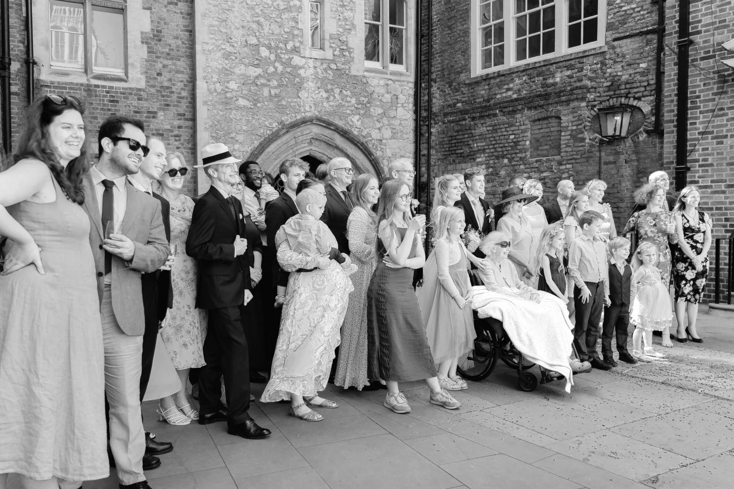 A large group of people, including men, women, and children of various ages, standing outside in front of a brick and stone building with large windows. They are smiling and appear to be attending a celebration or event.