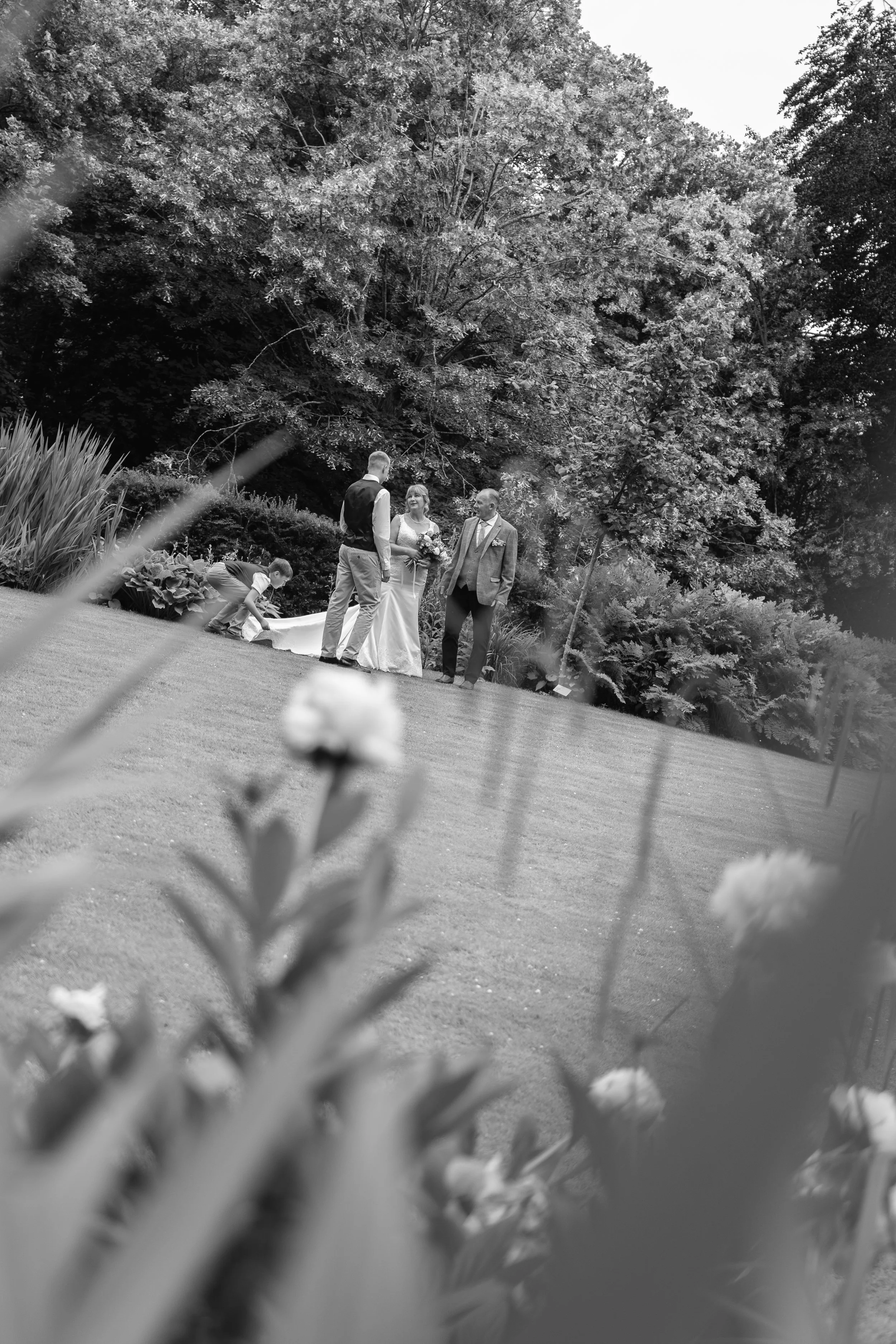 A black and white wedding scene outdoors with a bride in a white dress holding a bouquet, a groom in a light-colored suit, and another man in a jacket, standing on grass near a large tree, with a person crouching nearby and flowers in the foreground.