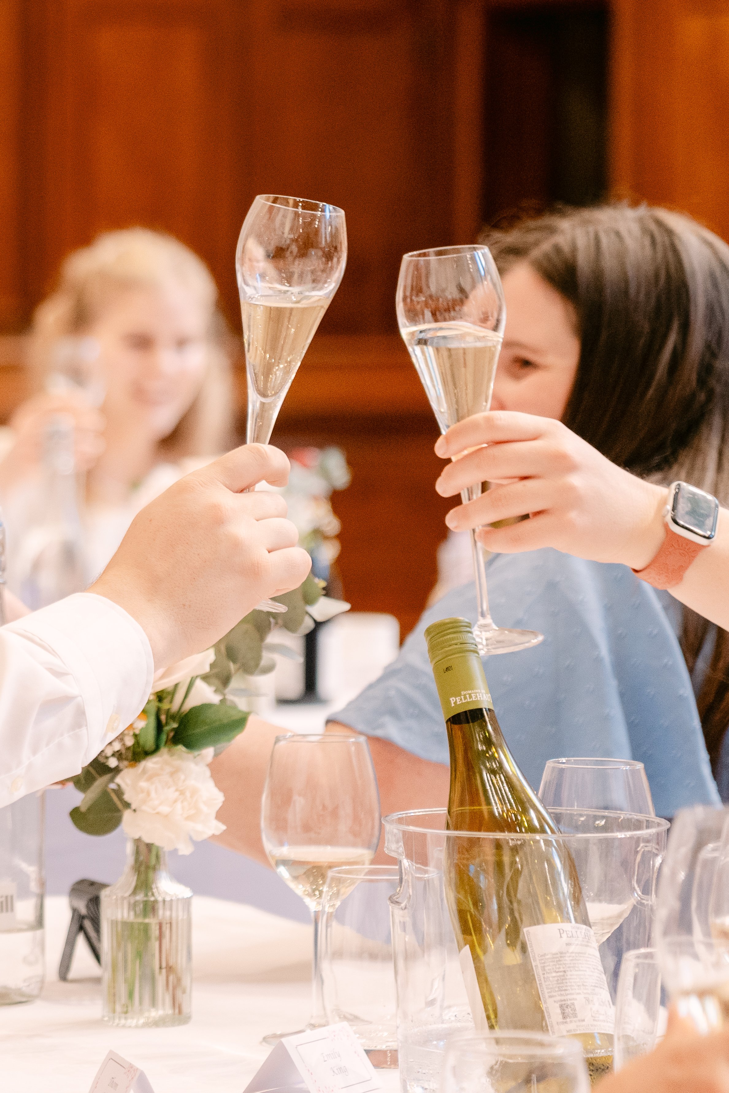 People celebrating with glasses of champagne at a dinner table, with a wine bottle in a bucket and floral arrangements visible.