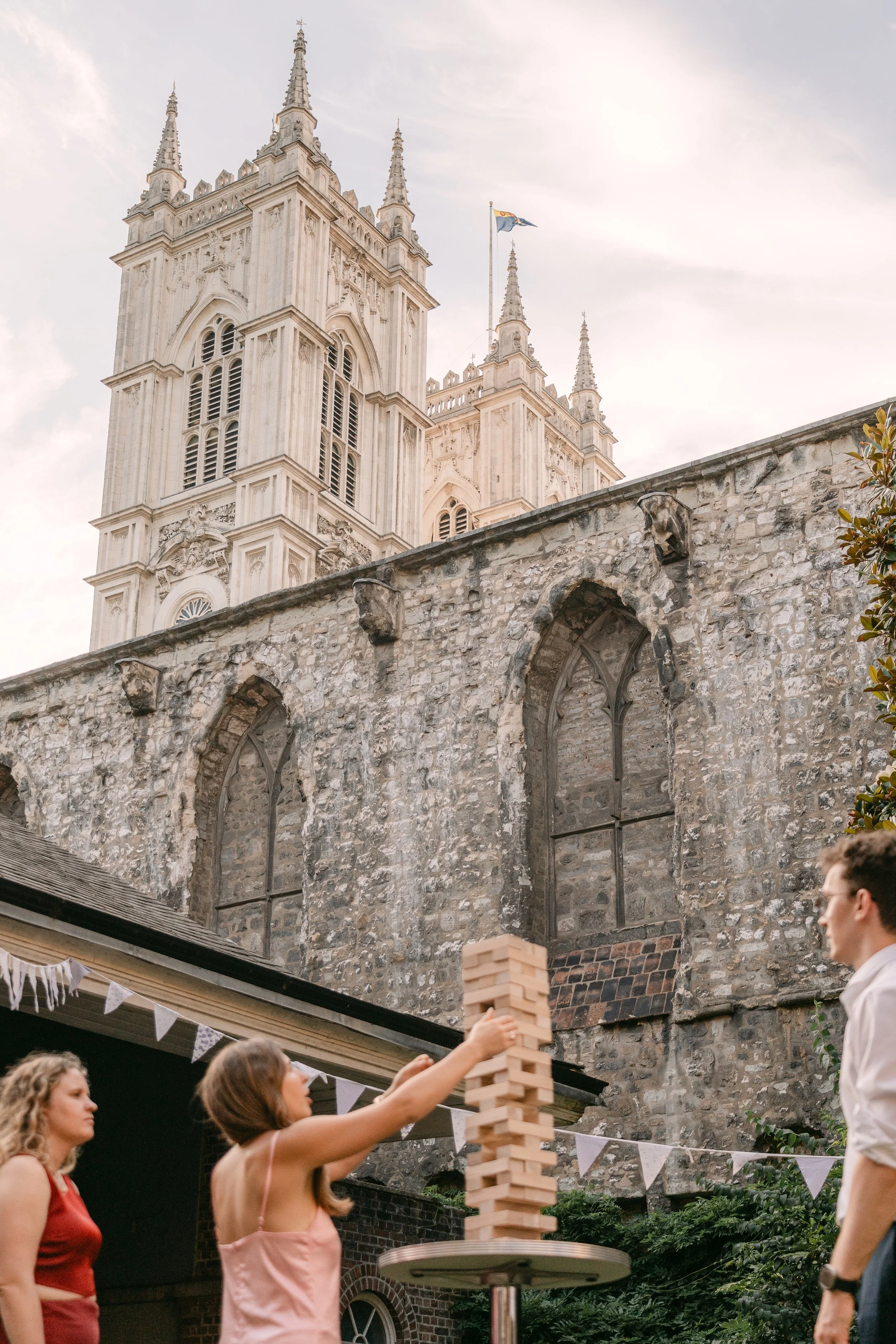 People playing Jenga in a courtyard with an old stone building, likely a church or cathedral, in the background, and a stack of wooden blocks on a round table.