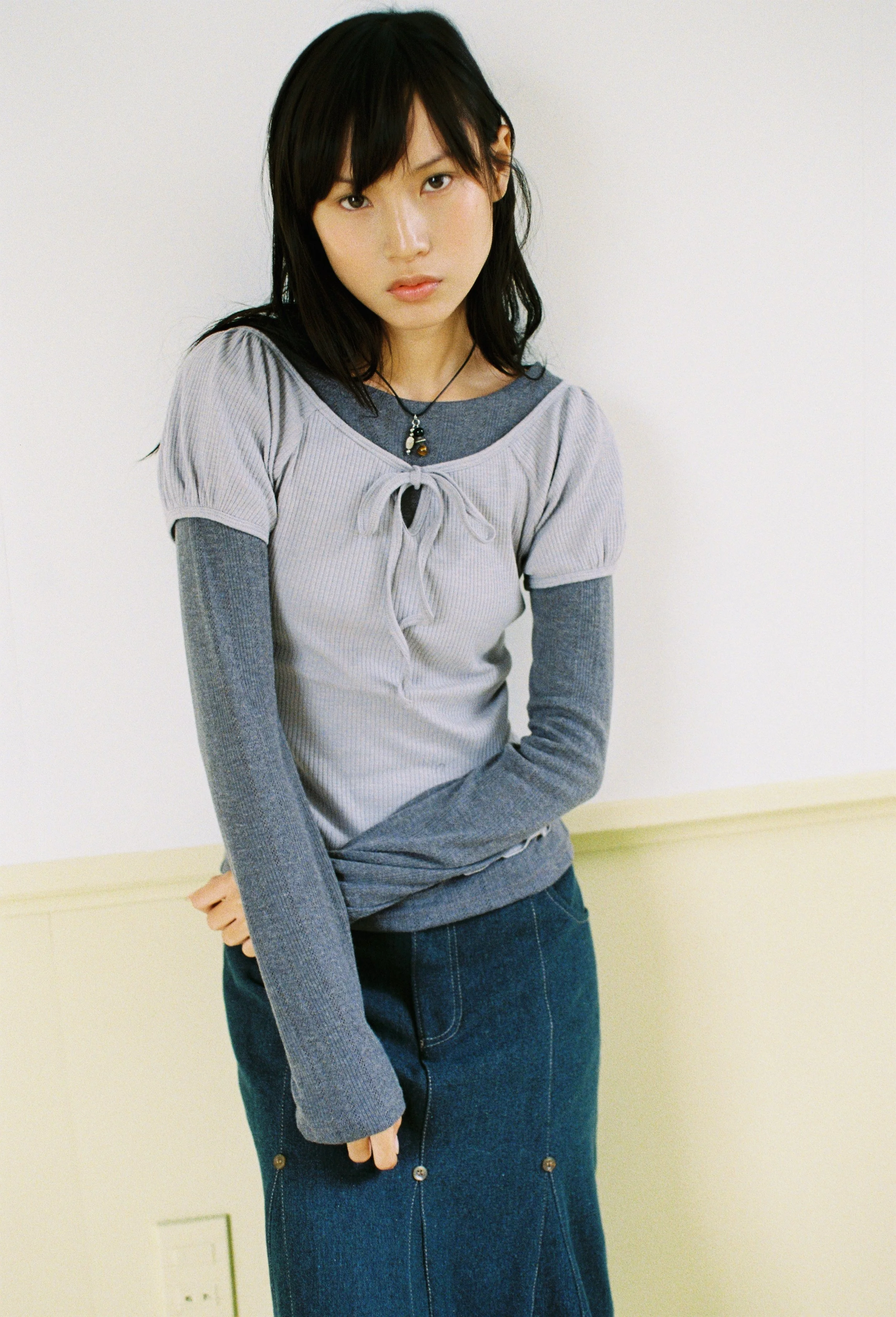 A young woman with black hair and fair skin, wearing a layered outfit of a gray short-sleeve top over a long-sleeve shirt and a denim skirt, standing against a plain light-colored wall.