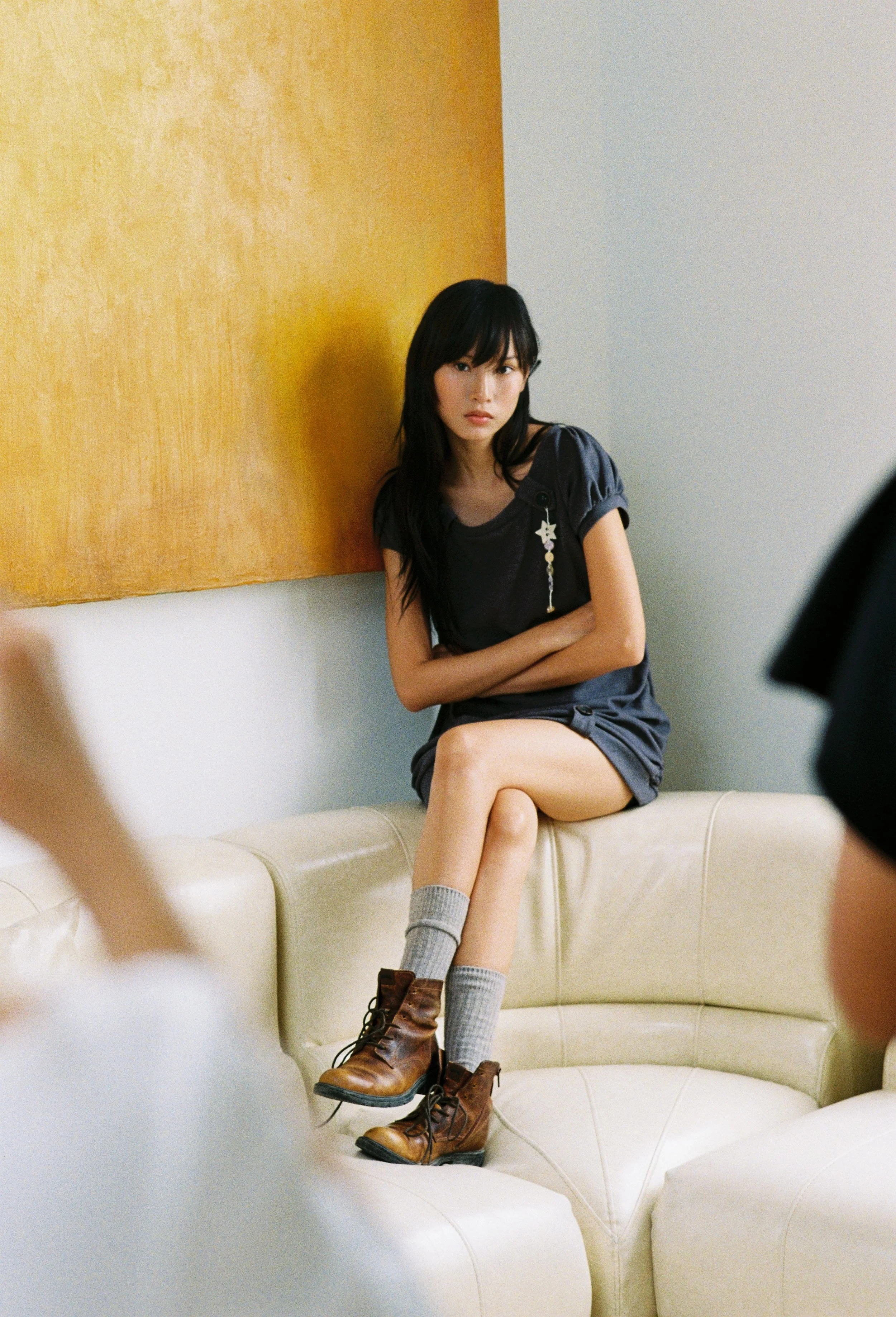 A young woman with long black hair, wearing a dark t-shirt, gray socks, and brown lace-up boots, sitting on a cream-colored, curved sofa with her arms crossed, looking to the side. She is positioned against a wall with a yellow panel and a light-colored wall.