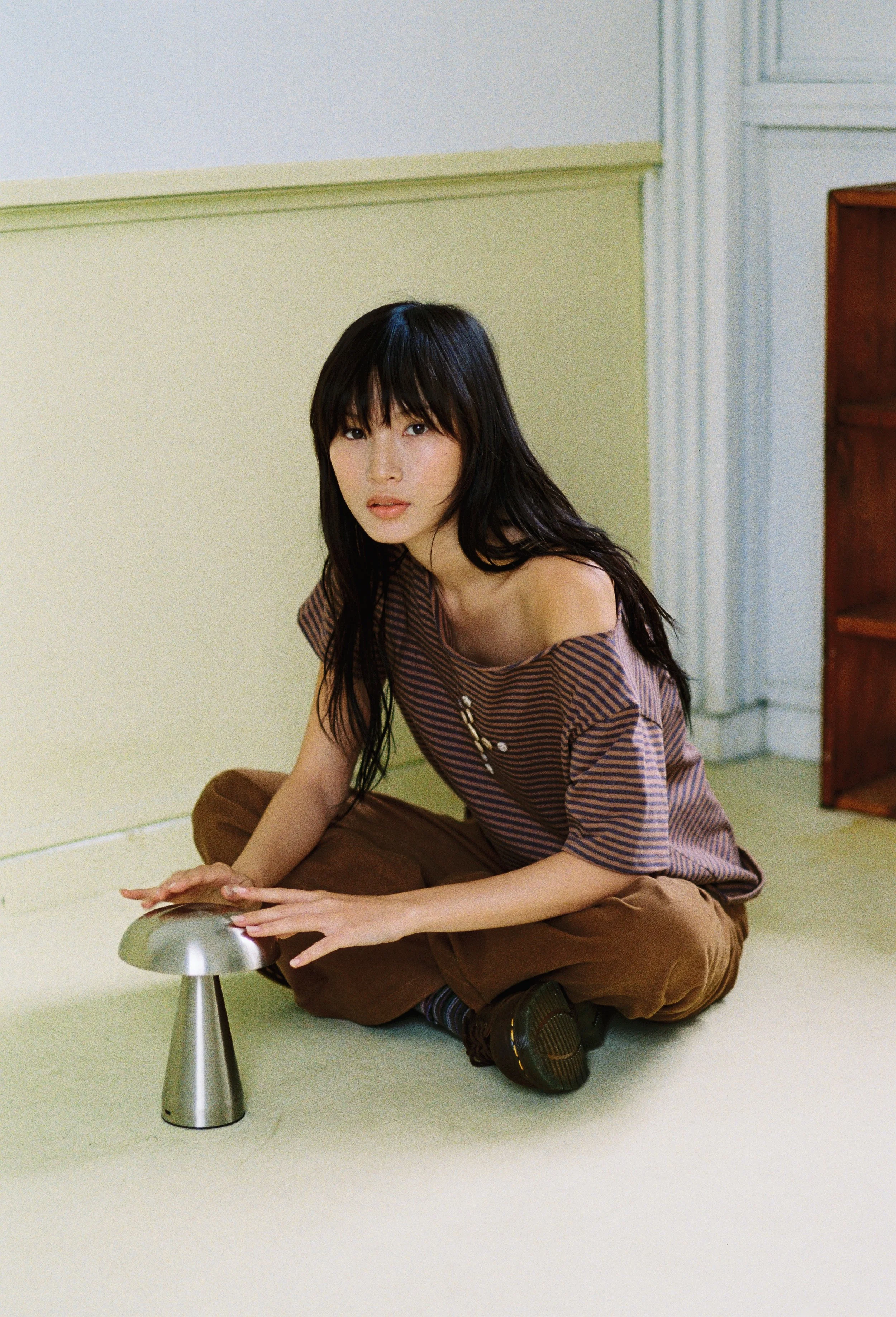 A young woman with black hair and bangs, wearing a striped shirt and brown pants, is sitting on the floor. She is holding a metallic object while looking at the camera. The setting appears to be a simple room with light-colored walls and minimal furniture.
