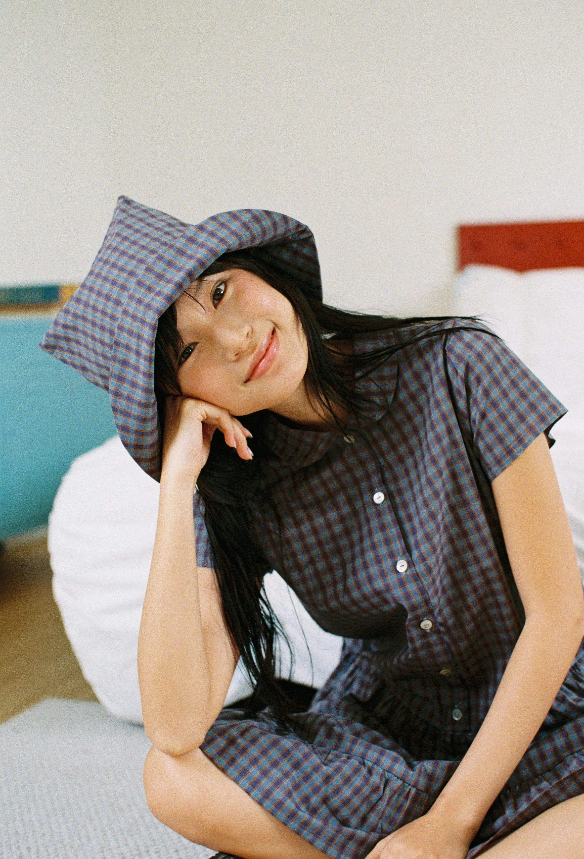 A young woman with long black hair, wearing a plaid shirt and a matching wide-brim hat, is sitting on the floor and smiling softly.