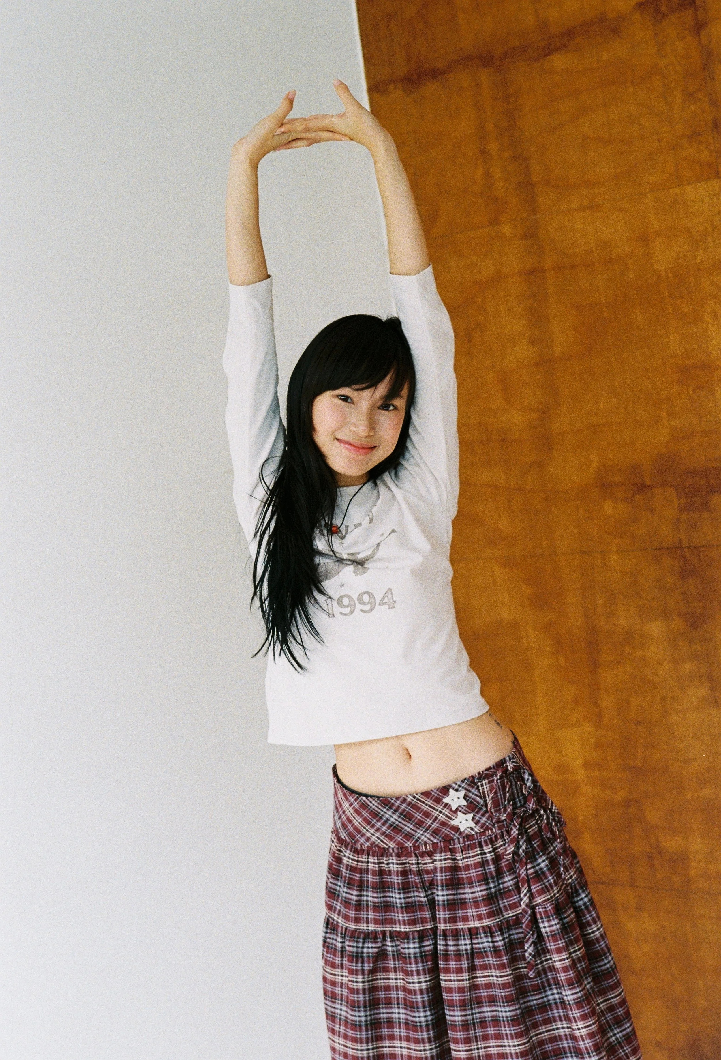A young Asian woman with long black hair is standing against a white wall with a wooden floor behind her. She is smiling and stretching her arms above her head, wearing a white long-sleeve shirt and a red plaid skirt.