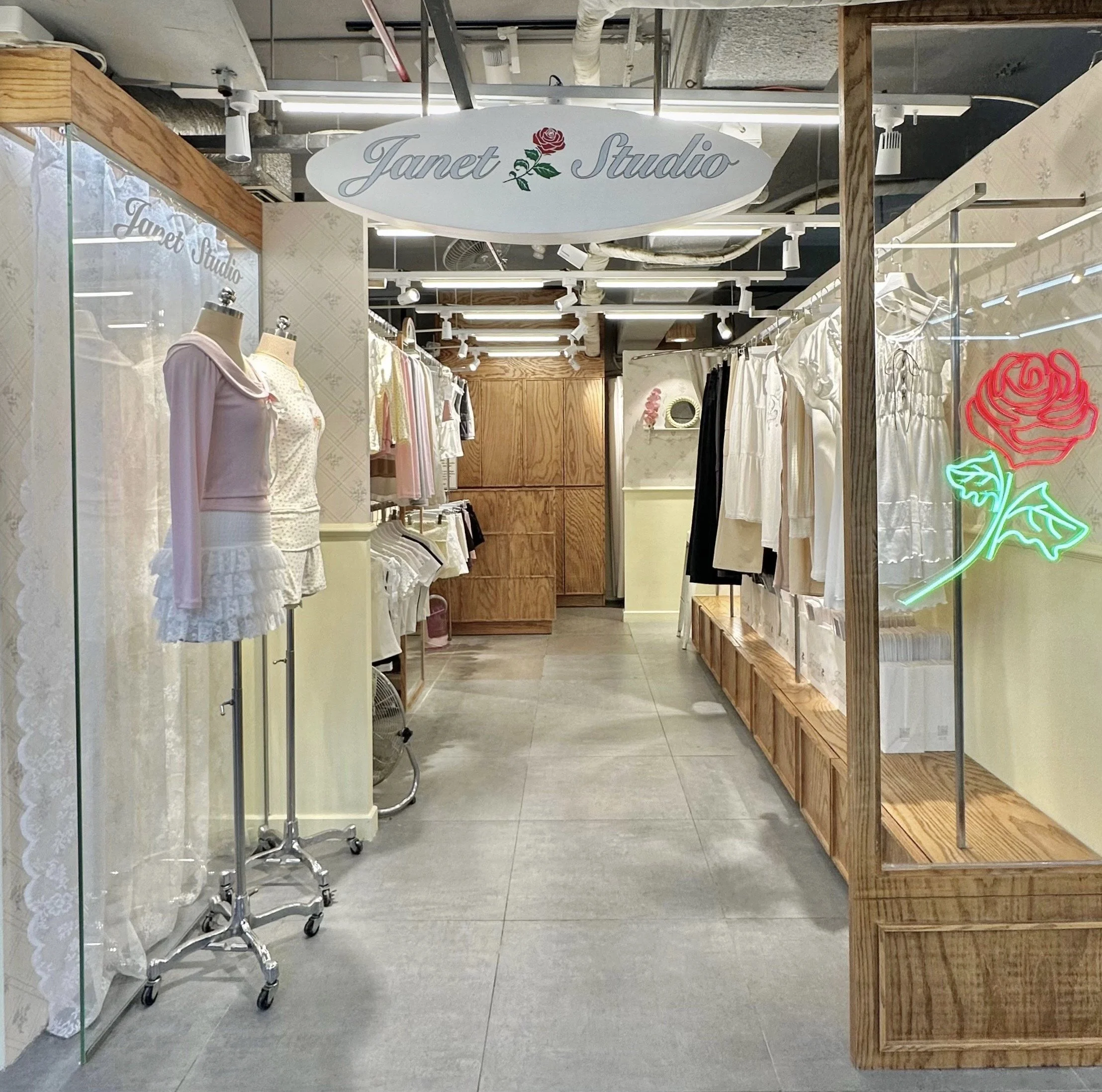 Store display with mannequins dressed in pastel pink and white sleepwear or loungewear, inside a shop named Janet Studio. The shop has glass display cases, wooden shelves, and features a neon light in the shape of a pink rose.