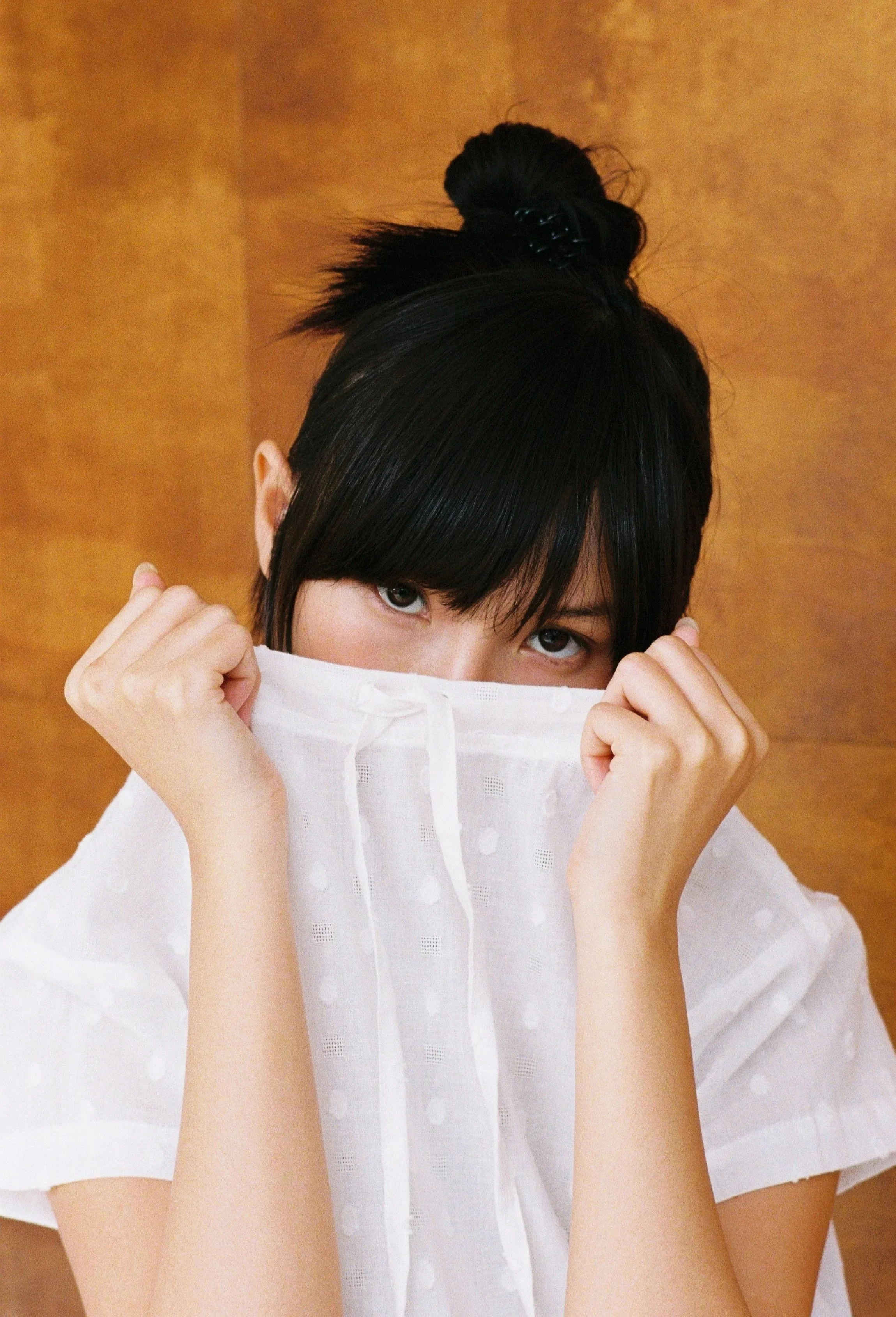 A person with dark hair tied in a bun, holding a white garment or mask in front of their face, against a warm brown background.