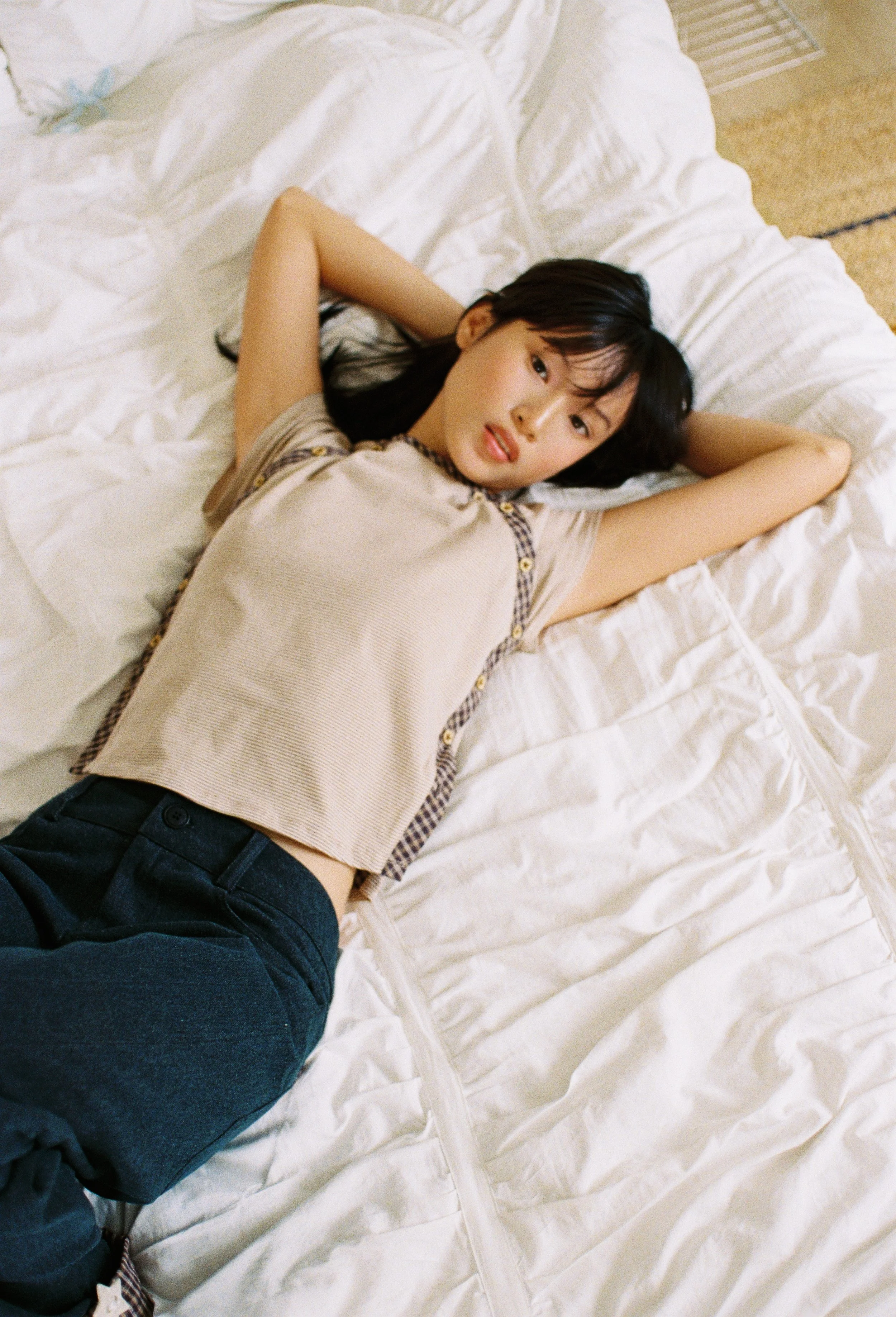 A young woman with black hair lies on her back on a bed with white sheets, looking at the camera with her arms bent behind her head. She is wearing a beige top with plaid trim and dark blue jeans.