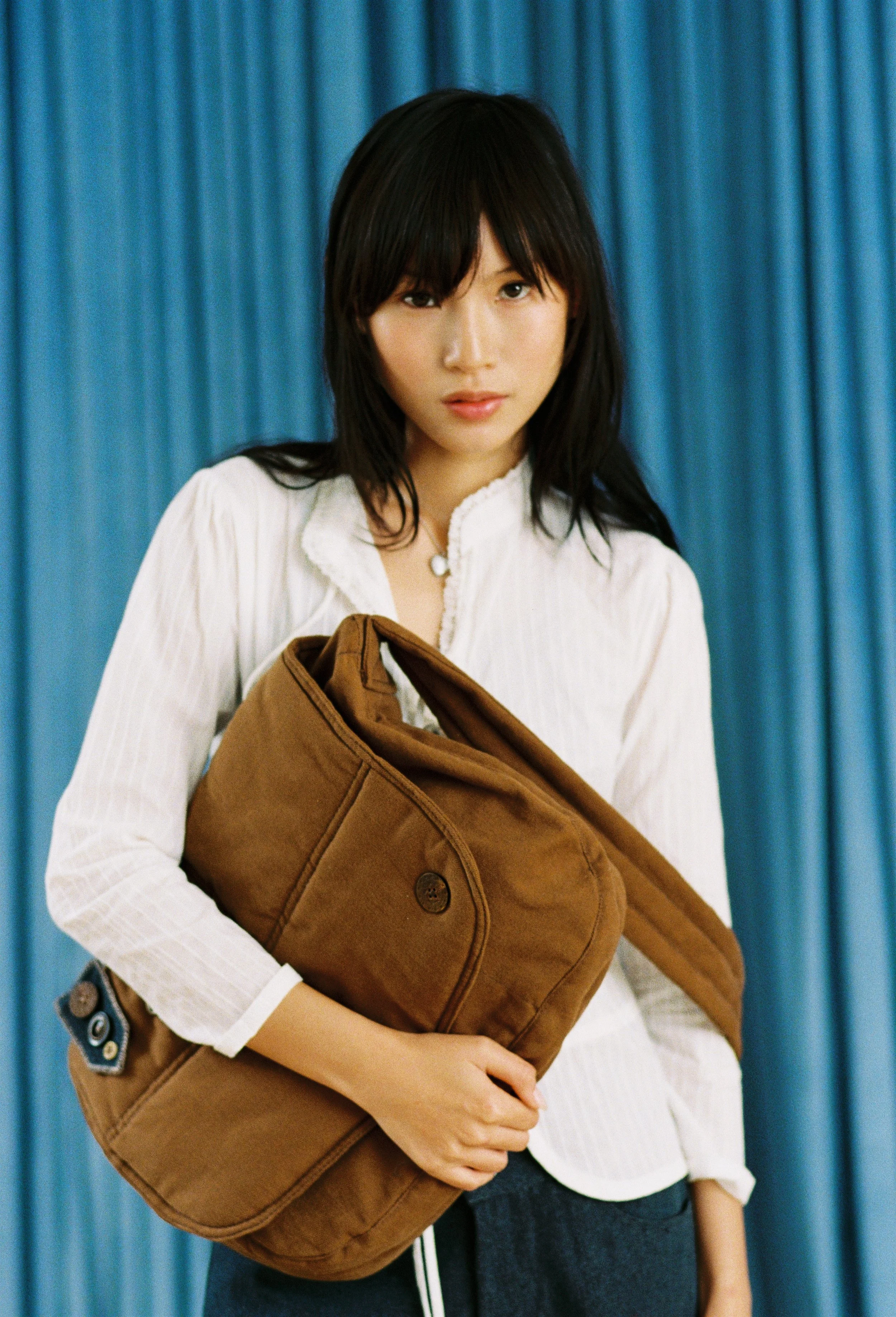 Young woman with dark hair, white blouse, and jeans holding a large brown bag in front of a blue curtain.