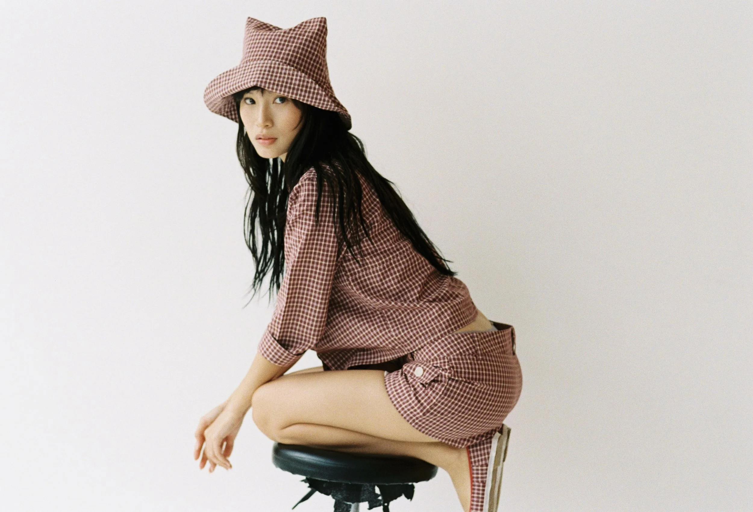 A woman with long black hair wearing a checkered dress and a matching hat, sitting on a stool against a plain white background.
