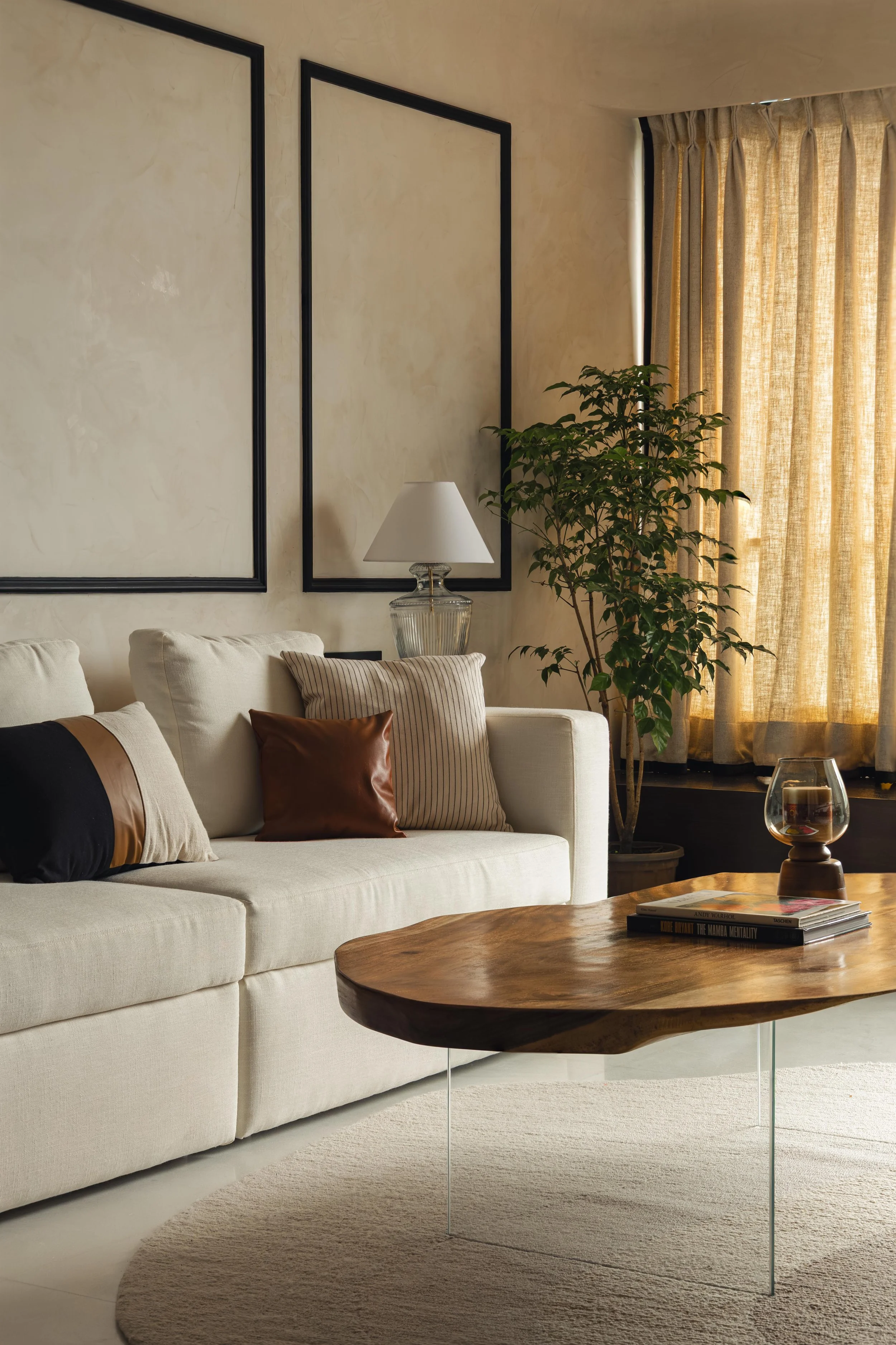 Living room with a white sofa, assorted pillows, a wooden coffee table, a large potted plant, a table lamp, and beige curtains covering a window.