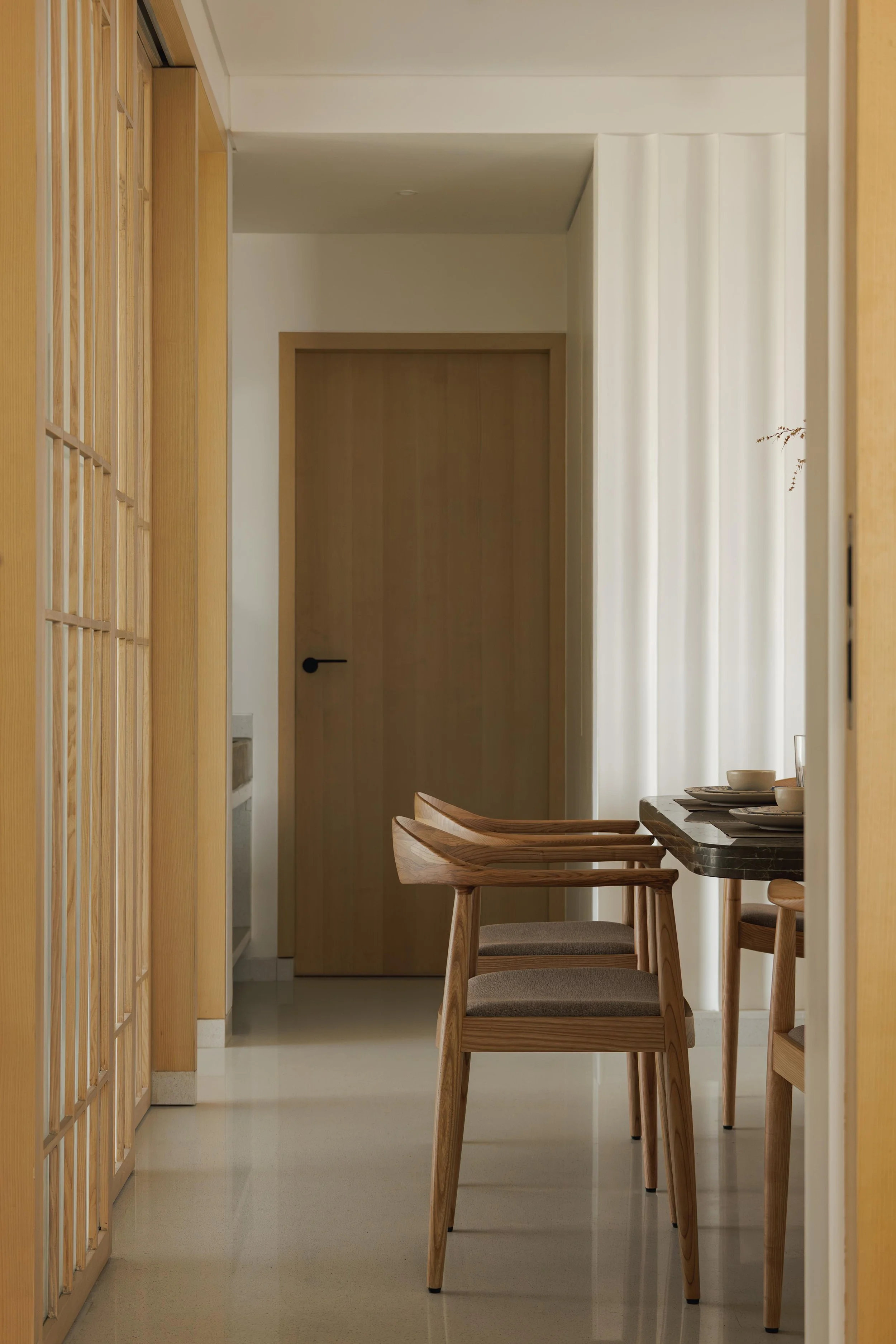 Interior view of a dining area with wooden chairs and table, cream-colored walls, a wooden door, and sheer white curtains, with natural light.