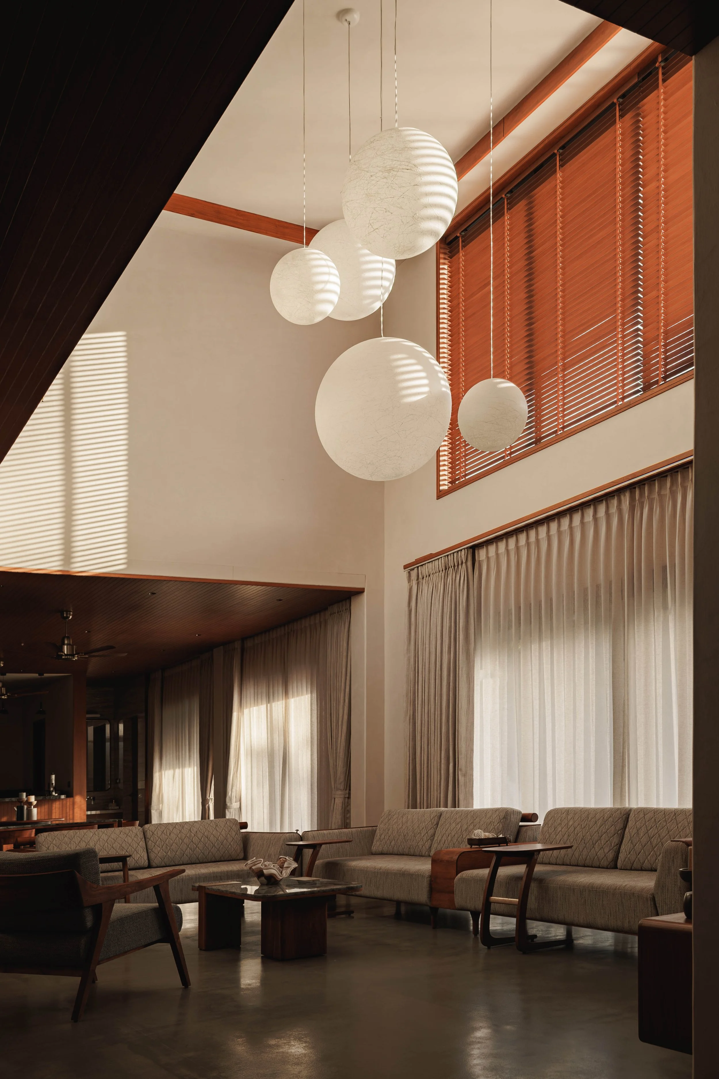 Living room with beige sofa, dark wood chairs, coffee table, large windows with curtains, hanging spherical lamp lights, and wooden blinds.