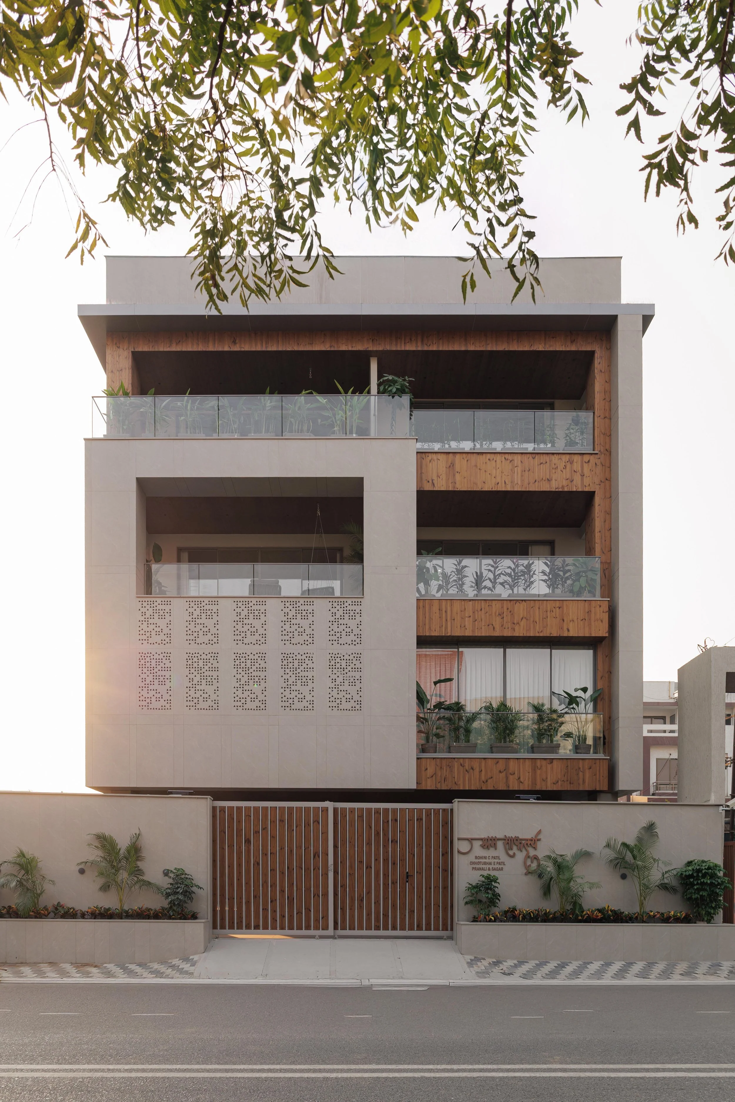 Front view of a modern three-story building with balconies, glass railings, and wooden accents, surrounded by small trees and plants, with a gated entrance at the street level.