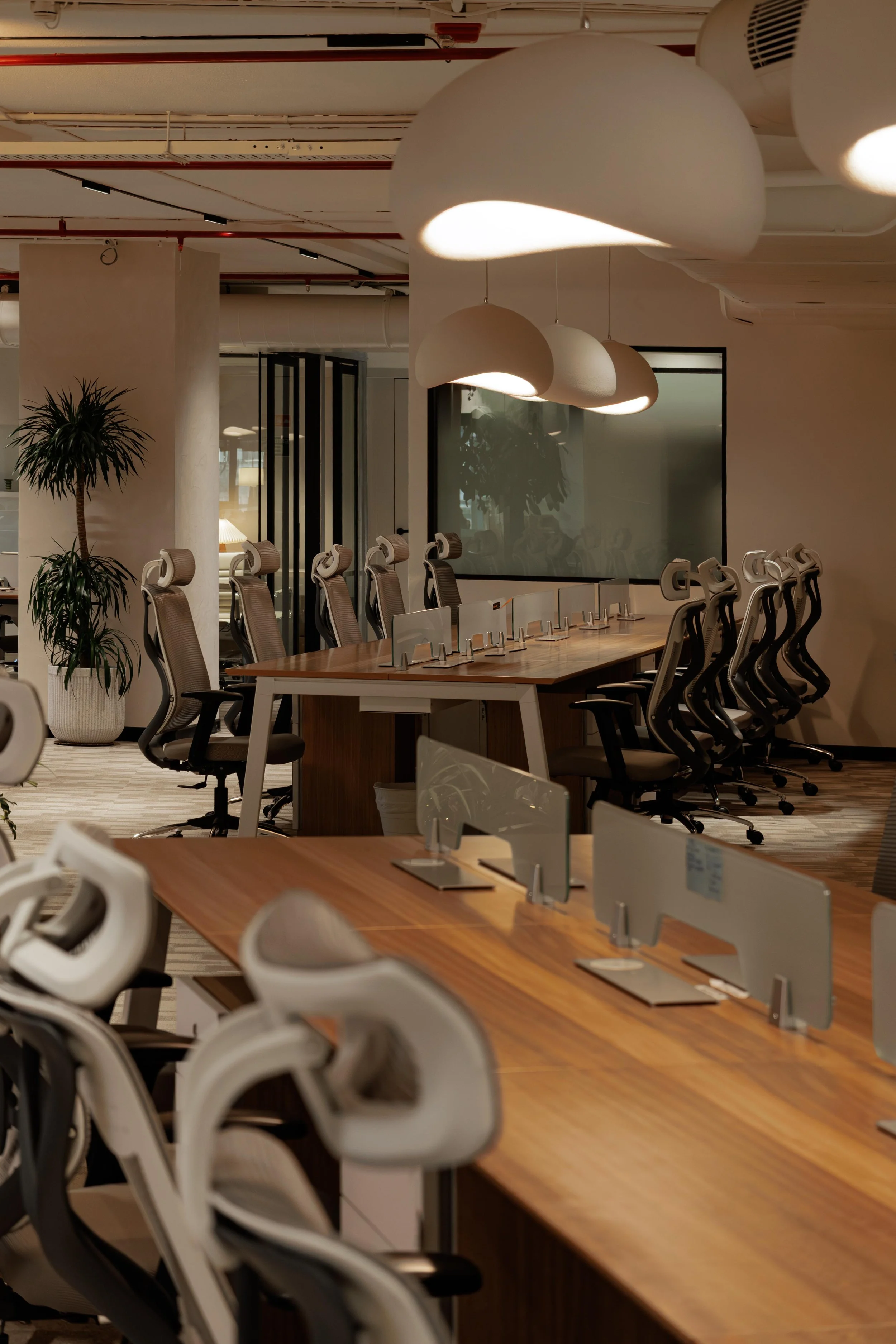 A modern conference room with a long wooden table and ergonomic chairs. There are hanging white pendant lights, potted plants, and a large window with reflective glass.