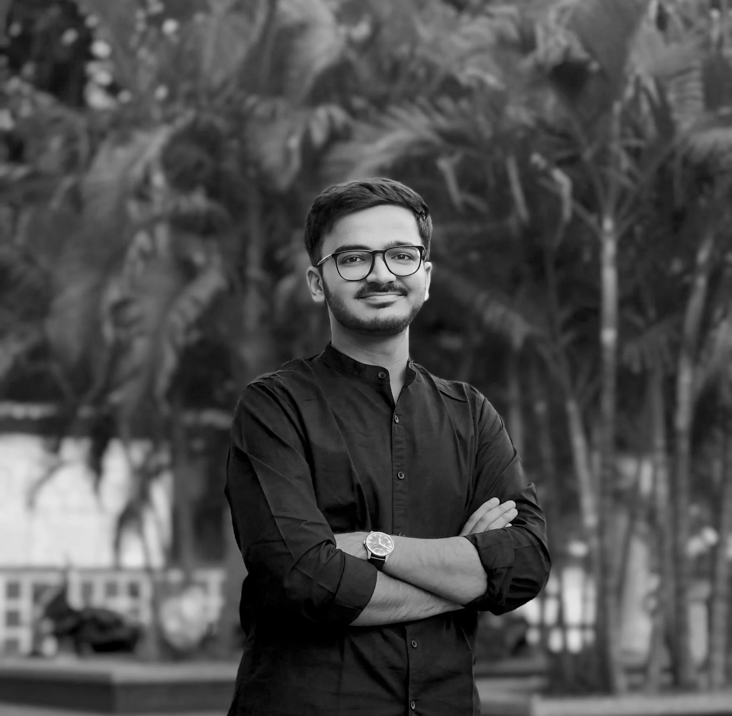 Black and white photo of the photographer with glasses, short hair, and a beard, standing outdoors with trees and plants in the background, wearing a dark button-up shirt with his arms crossed and a watch on his left wrist.