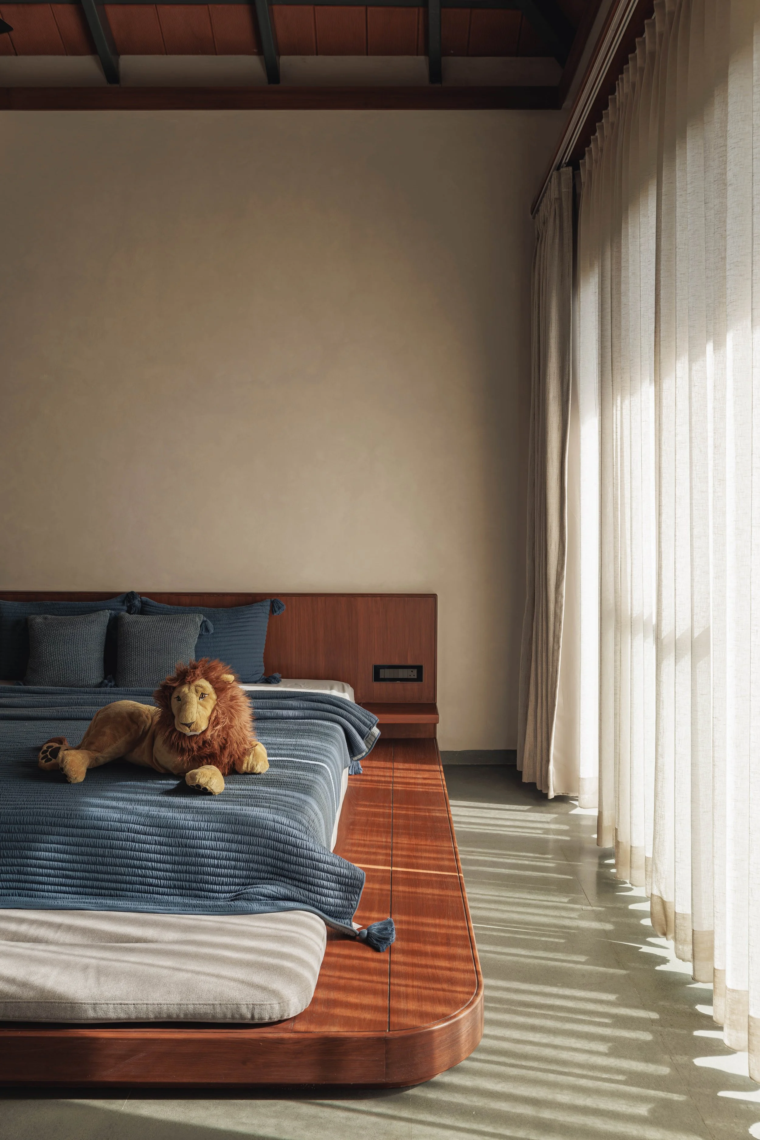 A bedroom with a bed covered in a striped blue and white blanket, topped with a plush lion toy, and surrounded by fluffy pillows. The room has a wooden headboard, large window with sheer white curtains, and sunlight casting shadows on the floor.