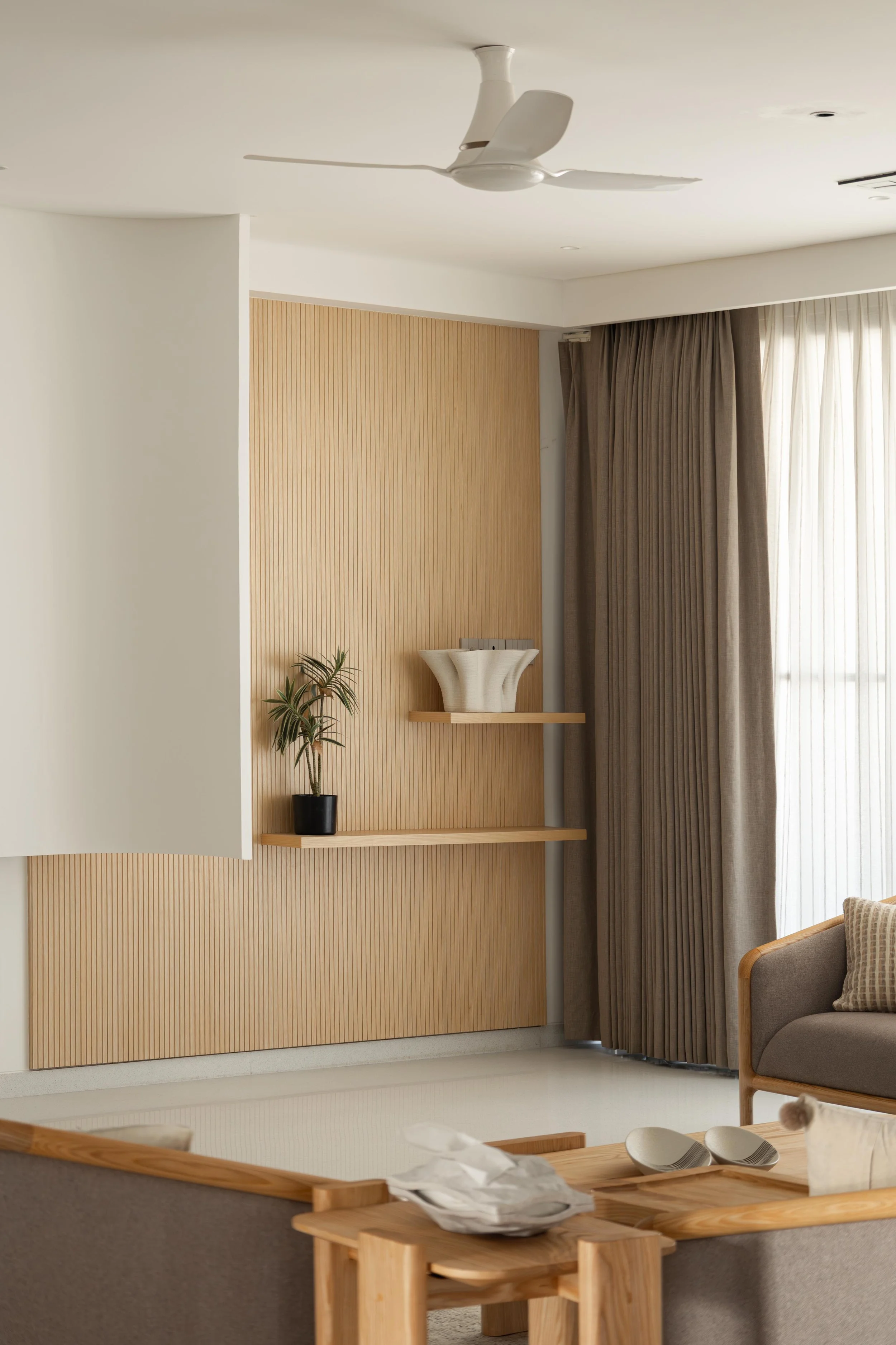 Interior of a modern living space featuring a wooden wall with vertical slats, beige curtains on a large window, a white decorative vase on a wooden shelf, a black plant pot with a green plant, and a cozy armchair with a cushion. Part of a wooden din
