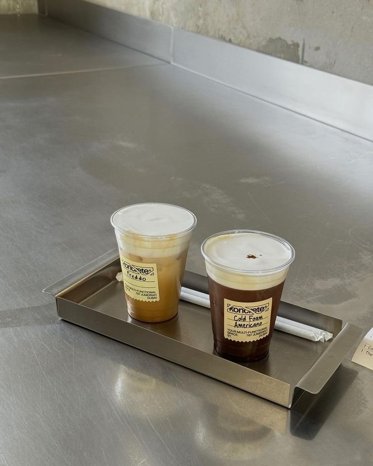 Deux verres de café, un froid et un chaud, sur un plateau en métal, posés sur un comptoir en métal avec fond en béton.
