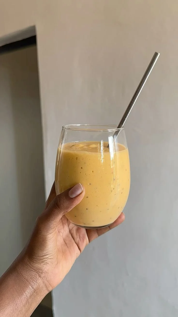 Une main tient un verre transparent rempli d'une boisson jaune, avec une paille en métal.