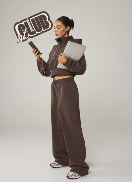 Une jeune femme porte un sweat à capuche marron, un pantalon ample assorti, et des baskets. Elle tient un téléphone portable dans une main, un ordinateur portable sous le bras, et porte des écouteurs. Il y a un logo « on CLUB » en arrière-plan.