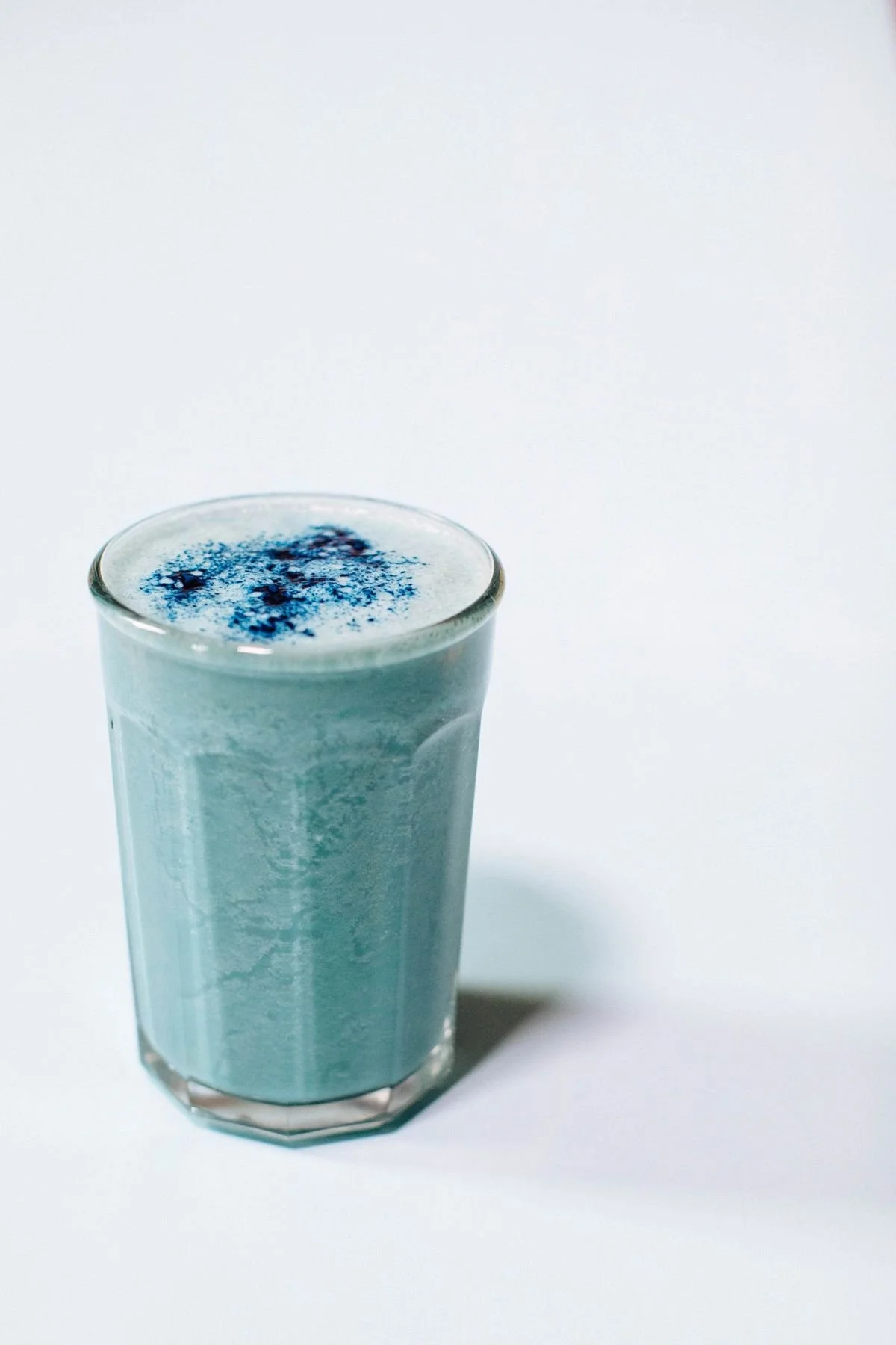 Un verre transparent rempli d'une boisson bleue avec des poudres bleues sur le dessus, posé sur une surface blanche.