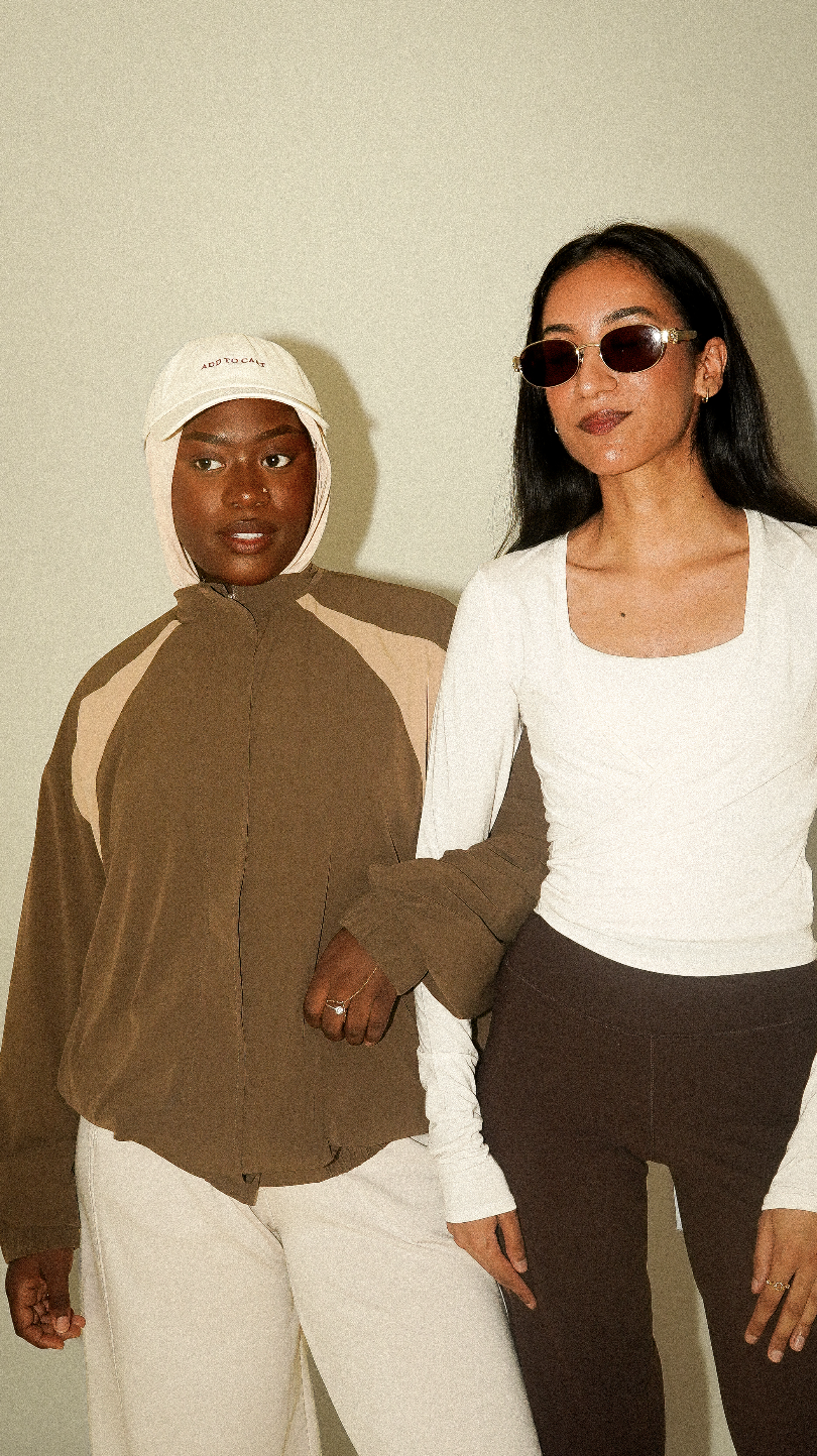 Deux femmes posent côte à côte, portant des vêtements de mode et des accessoires, dont des lunettes de soleil et un chapeau.
