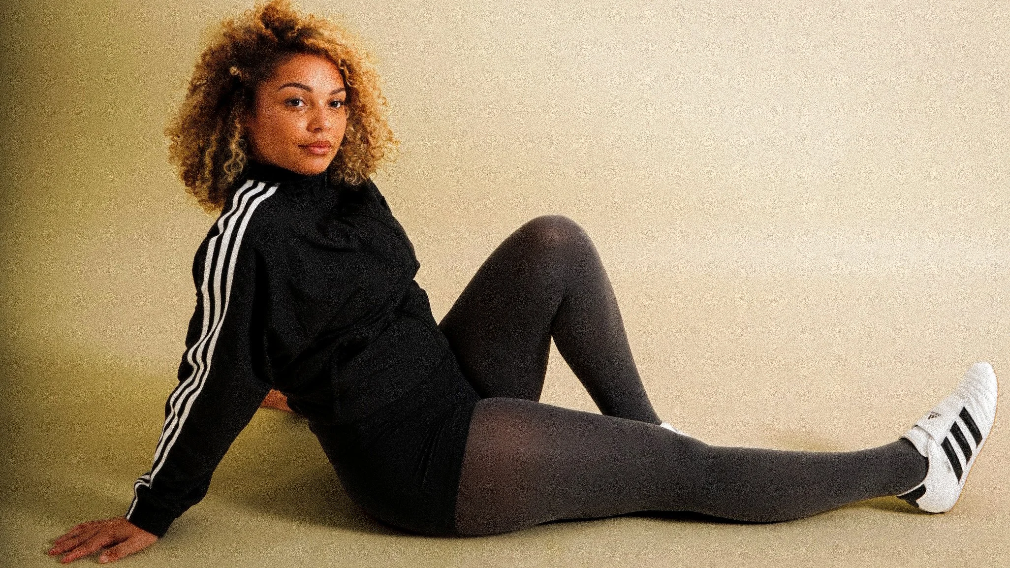 Jeune femme avec cheveux bouclés blonds, en tenue de sport noire avec bandes blanches, assise contre un mur beige, portant des chaussettes de sport Adidas à rayures vertes et noires.