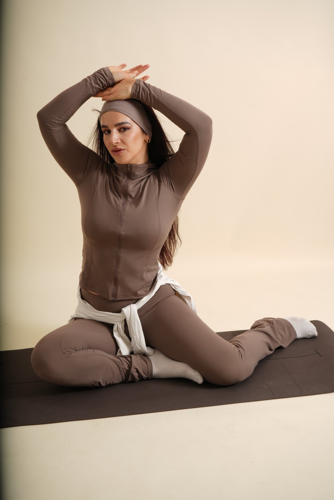 Femme en tenue de sport marron, faisant une pose de yoga sur un tapis, avec un bandeau à la tête et un sweat blanc noué à la taille.