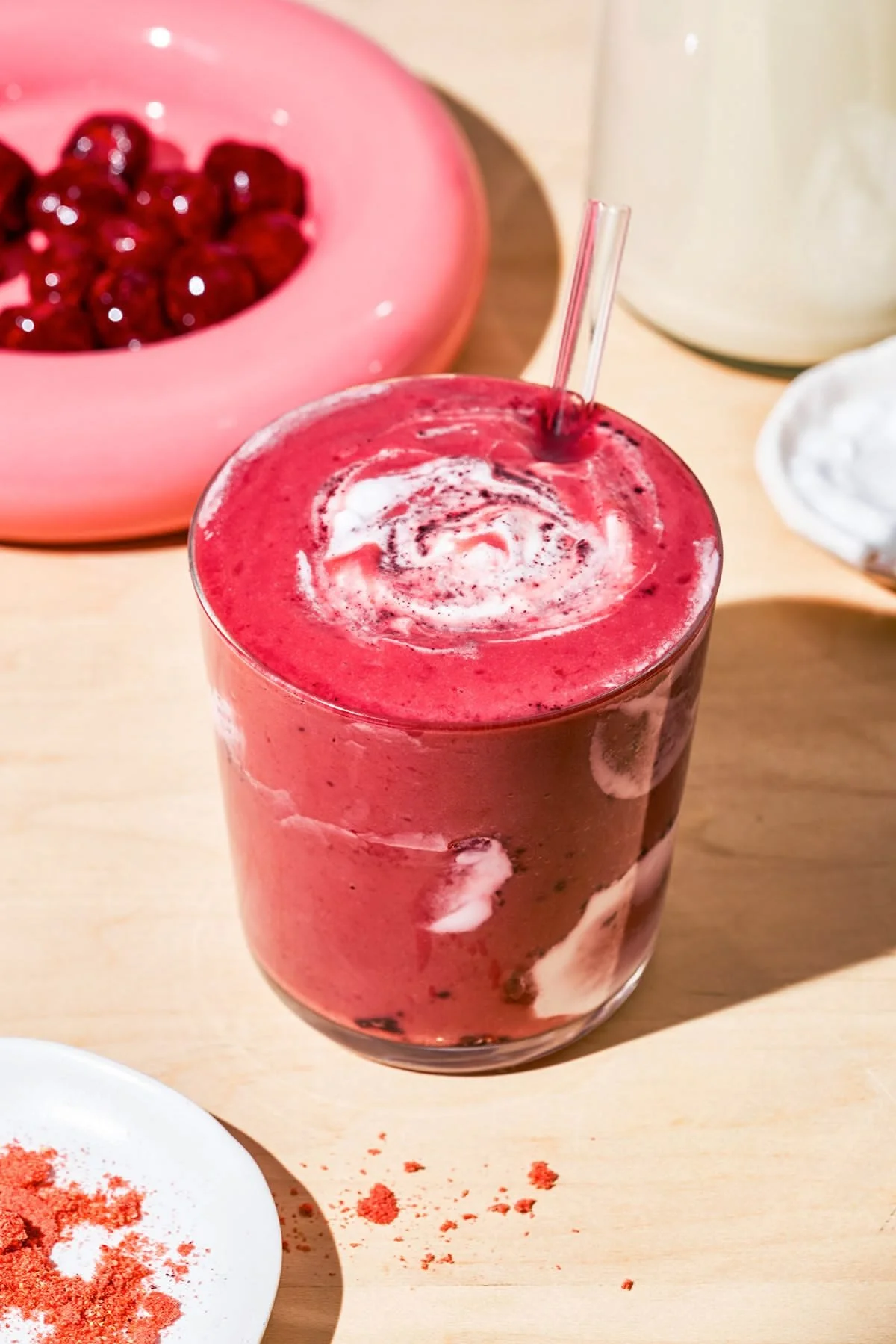 Un smoothie rose avec un bâtonnet en verre, entouré de fruits rouges et de poudres colorées sur une table en bois.