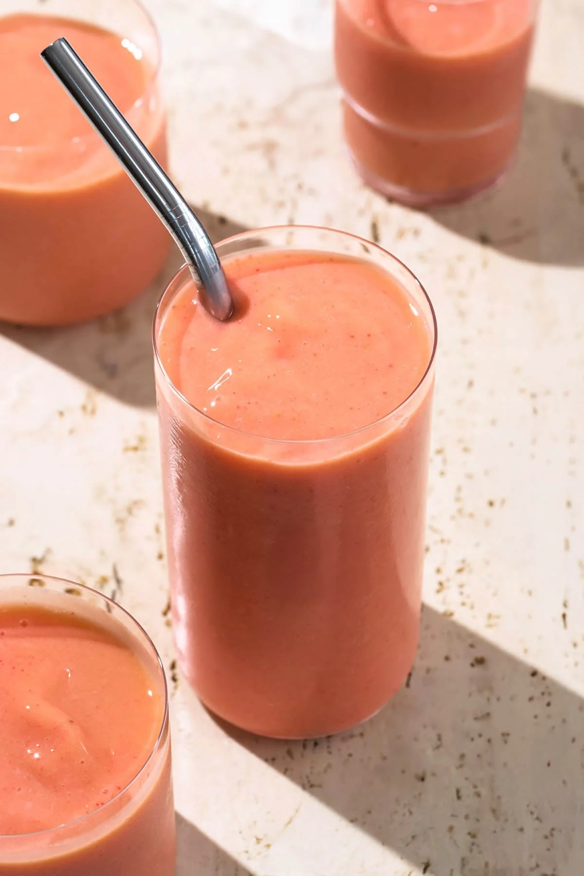 Verre de smoothie à la fraise avec une paille en métal, plusieurs autres verres de smoothies à la fraise en arrière-plan sur une surface beige.