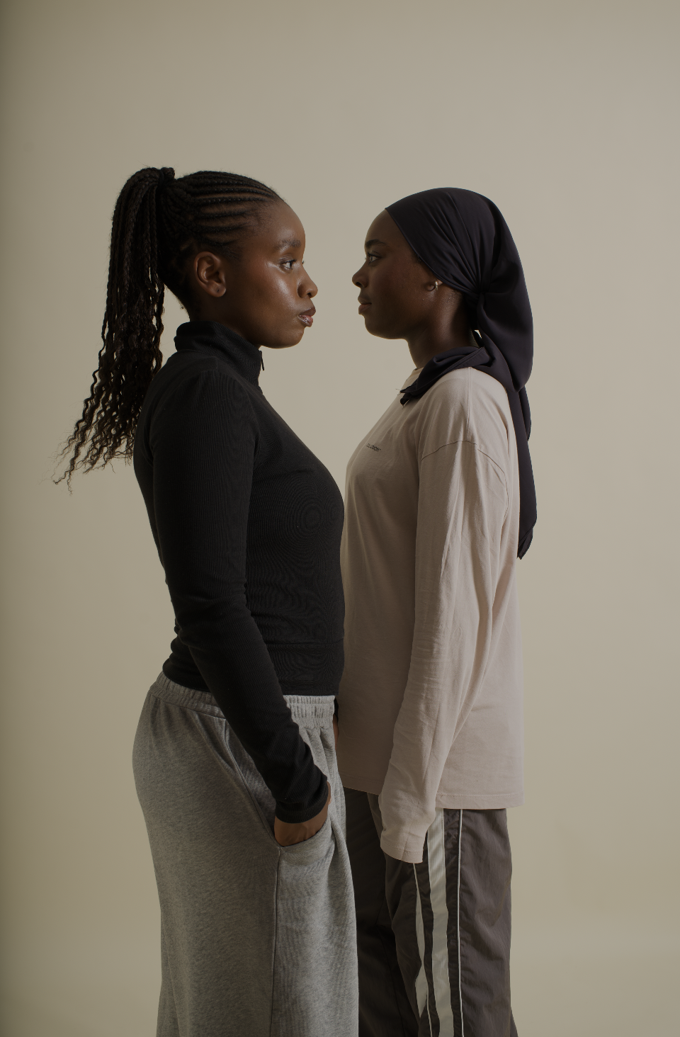 Deux femmes debout face à face, regardant intensément, en vêtements décontractés, dans un fond neutre.