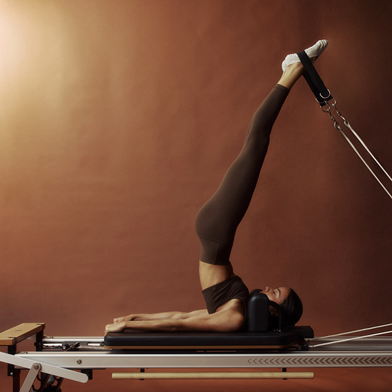 Femme faisant de la rééducation sur une machine Pilates, allongée, avec les jambes levées en l'air, les pieds dans des étriers, sur un fond marron.